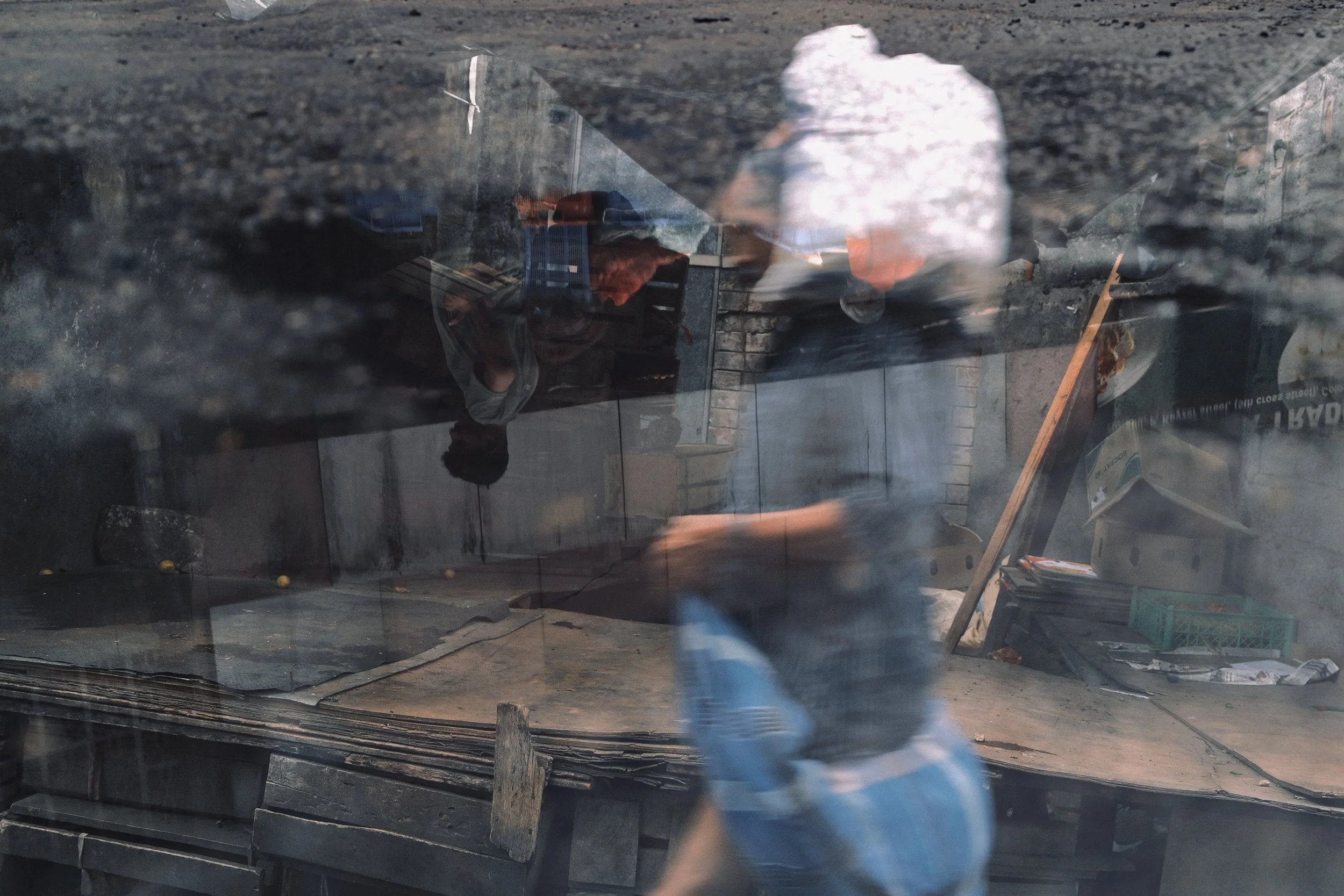 Reflection of two men through a glass window, with one man wearing a white shirt and the other in dark clothing, in an unfinished or cluttered space with boxes and wooden planks.