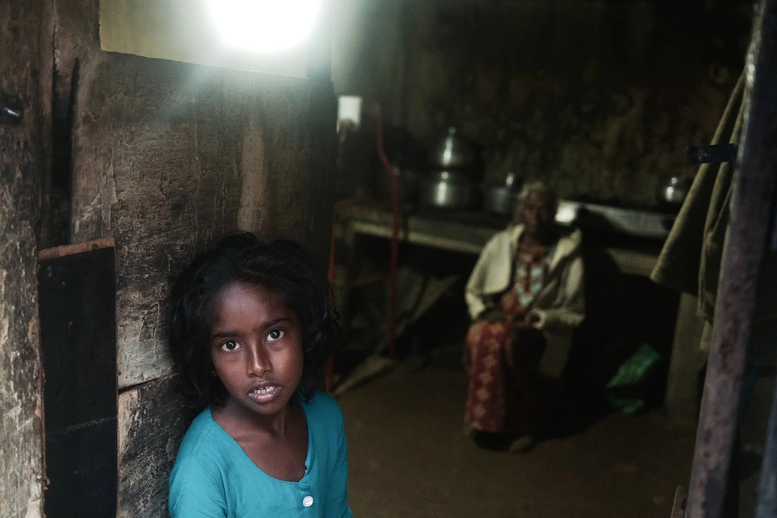 A young girl with dark hair and brown skin standing in a dimly lit, rustic room with a rough wall, looking directly at the camera. In the background, an elderly woman with dark skin and gray hair is seated, wearing a patterned skirt and a light-color