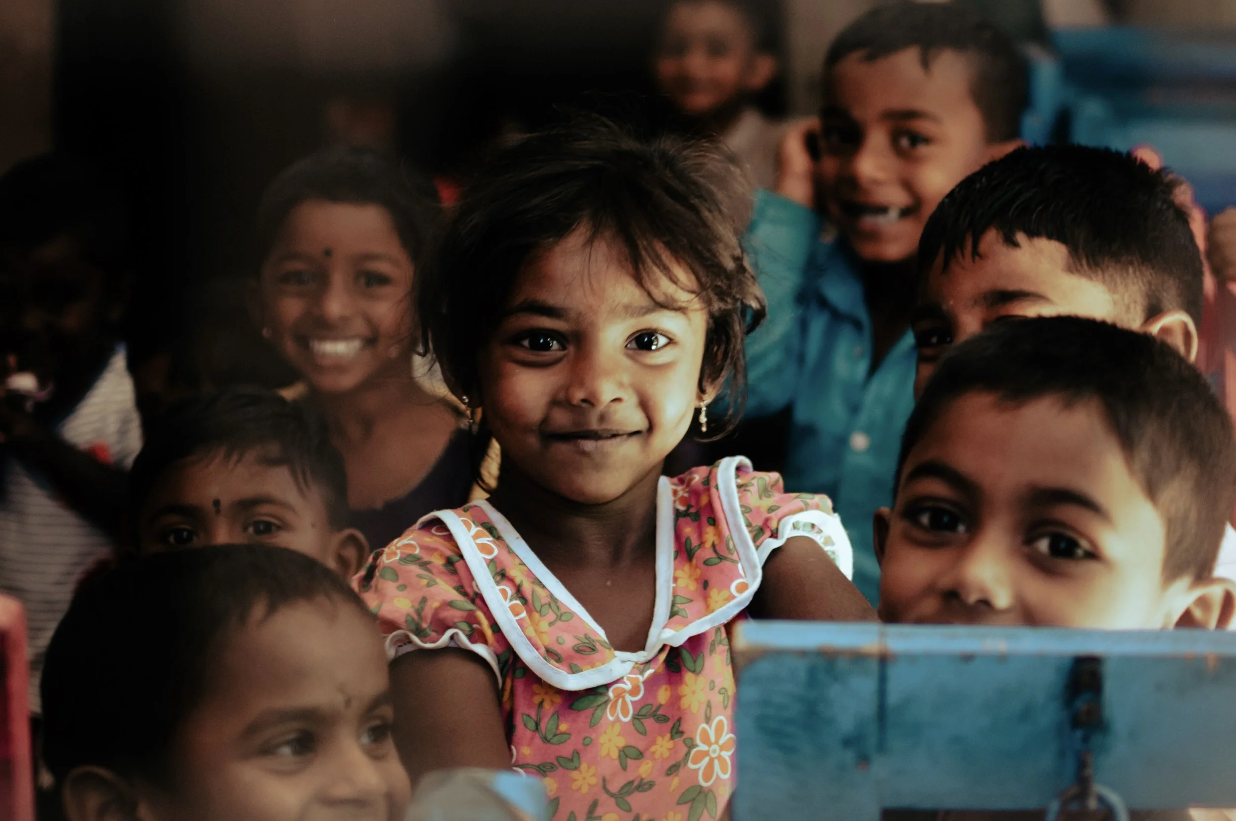Group of smiling children, with one girl in the center looking directly at the camera, wearing earrings and a floral dress with ruffled sleeves.