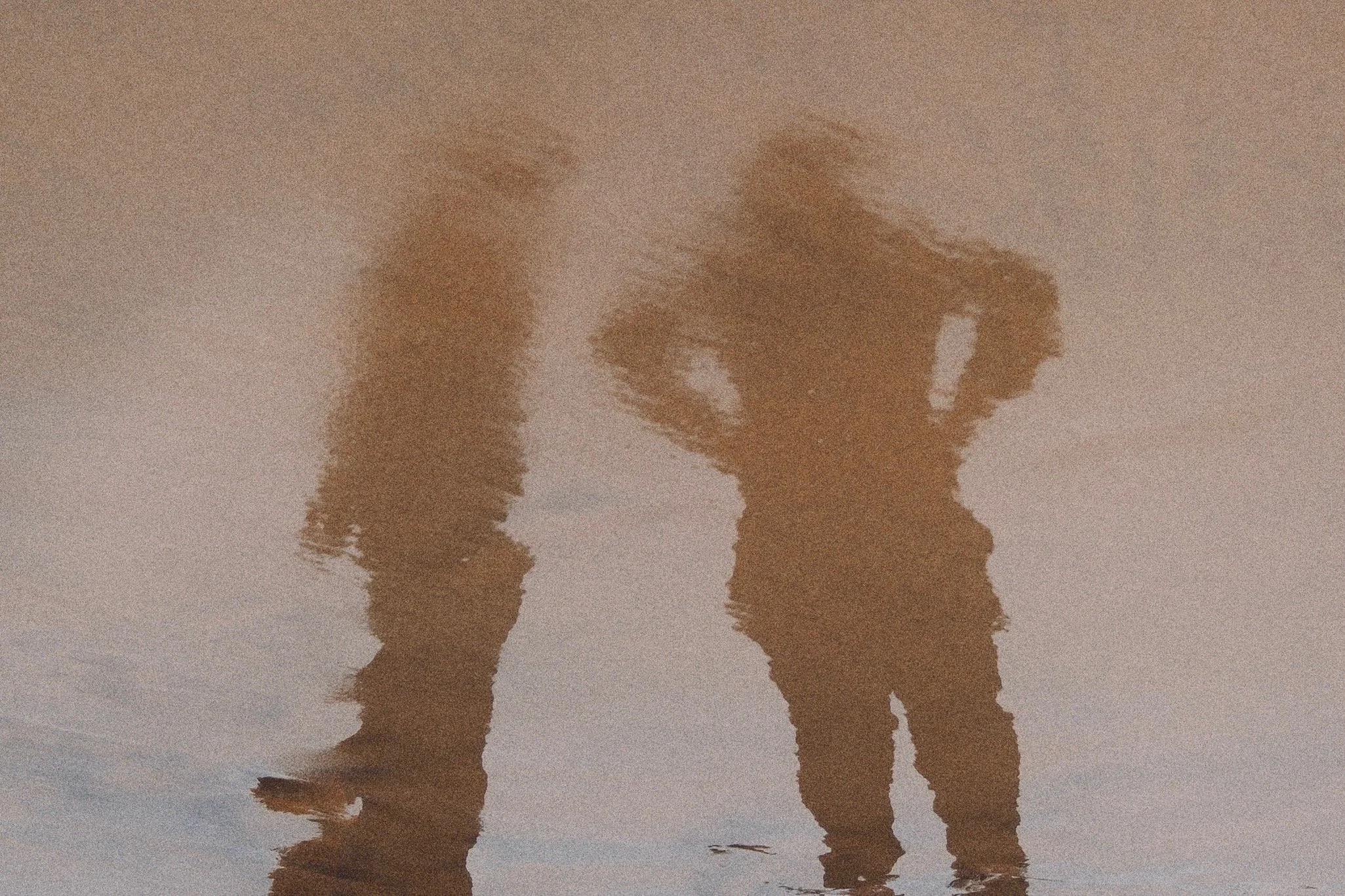 Reflection of two people walking on the beach at sunset, with their silhouettes visible in the wet sand.