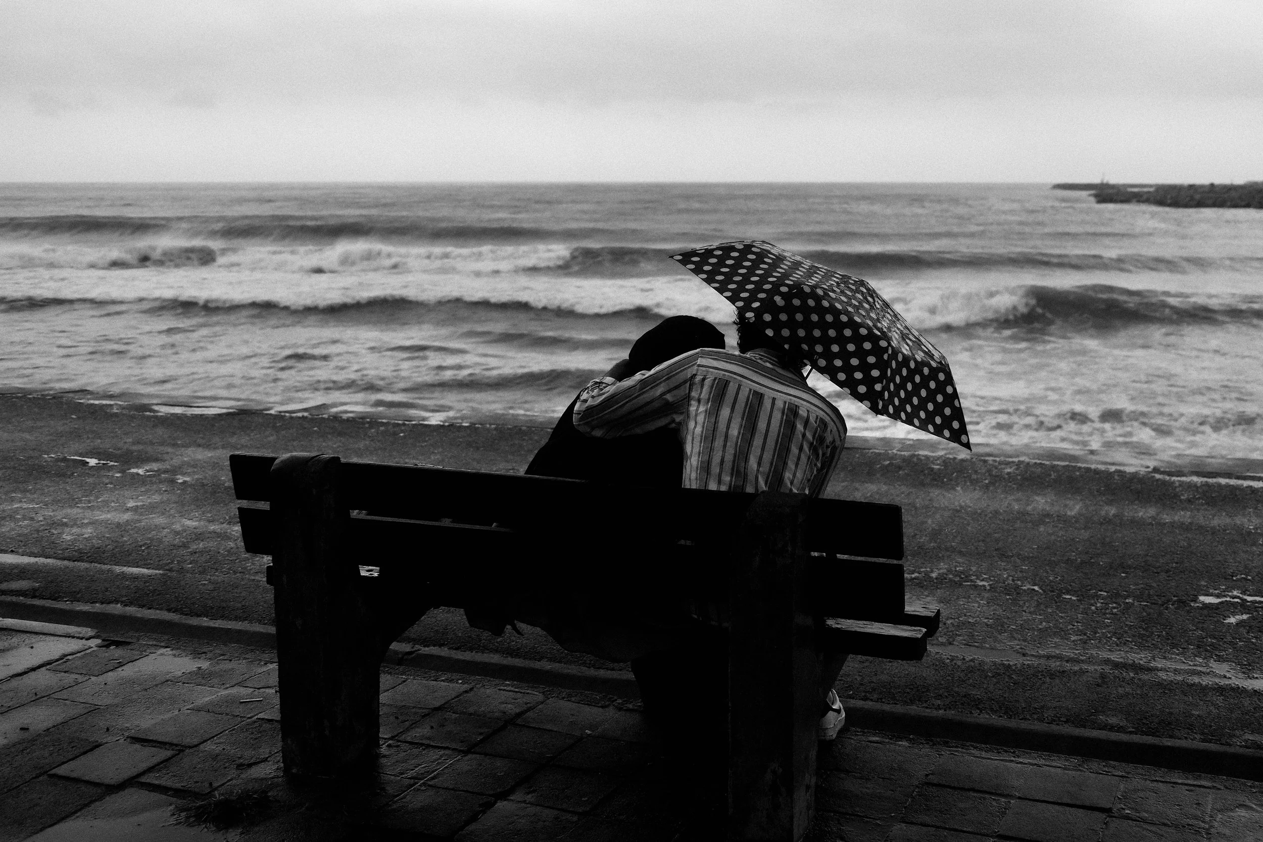 A black-and-white photo of two people sitting on a bench near the beach, with one person holding a polka dot umbrella. The ocean waves are visible in the background, and the scene appears to be overcast.