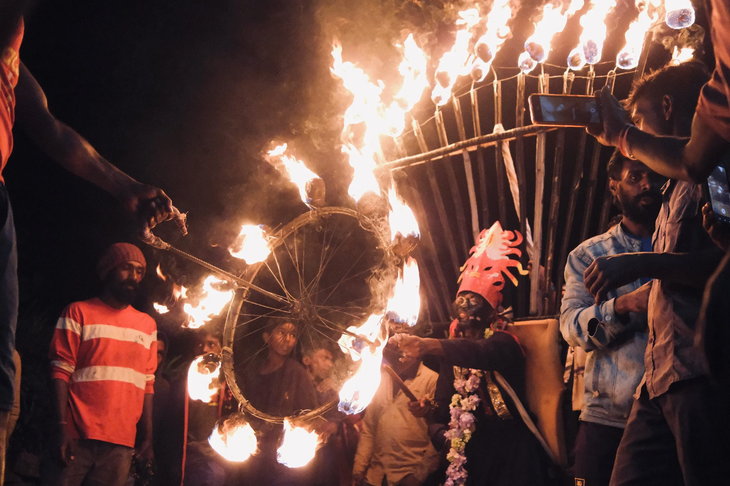 A traditional Indian or Hindu celebration involving fire and a decorated figure. People are gathered around, some taking photos, with one person holding a large flaming wheel with flames on its perimeter, and another person wearing a colorful headdress.