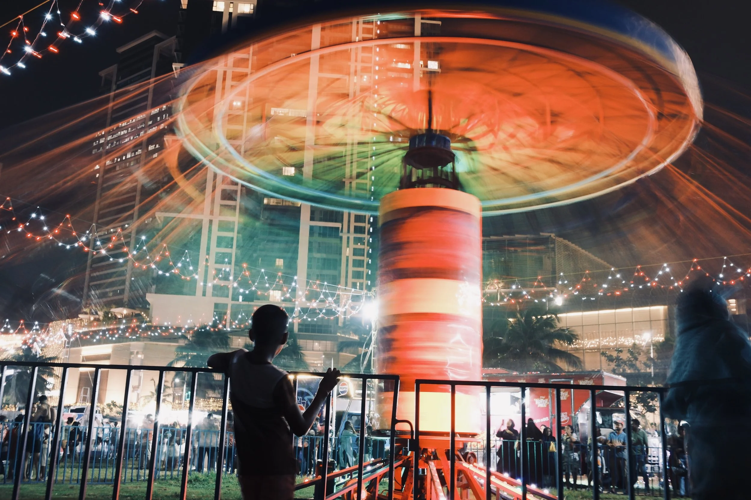Nighttime at a fair with a colorful spinning amusement ride creating motion blur, surrounded by people and string lights, in an urban setting with tall buildings.