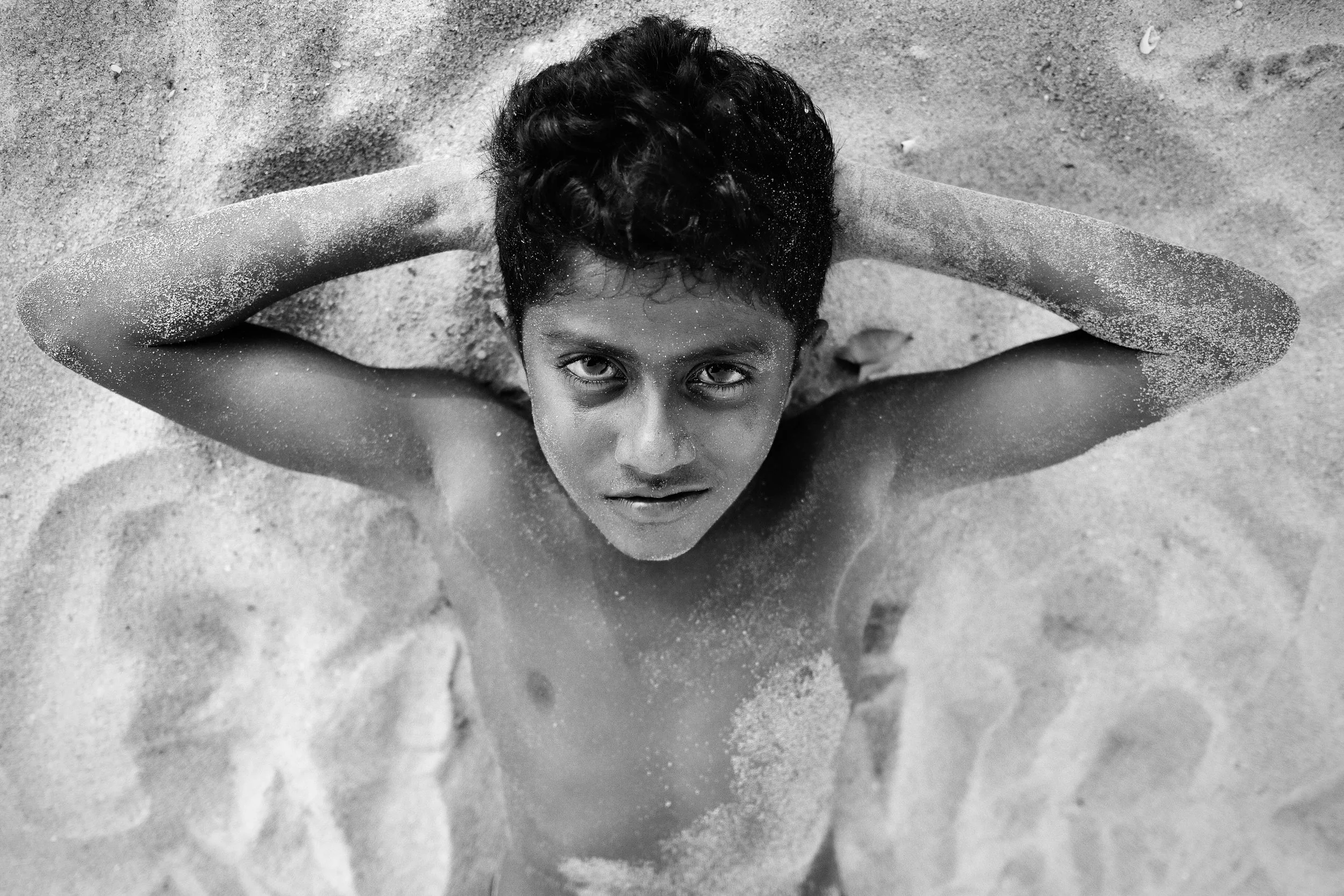A young boy with dark hair lying on sandy beach with arms behind his head, looking up at the camera in black and white.