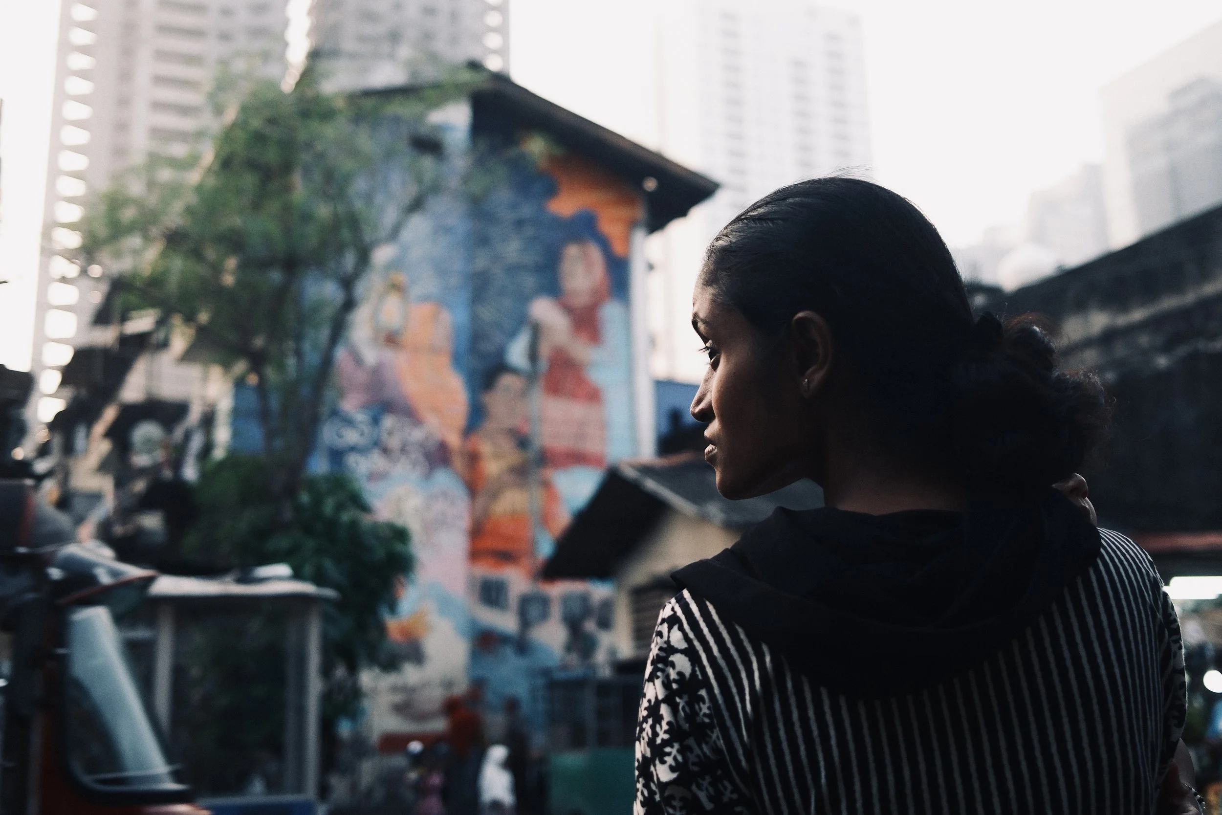 A woman with dark hair tied back, wearing a black and white striped shirt, standing outdoors in an urban setting, with a colorful mural of a woman in the background, surrounded by trees and tall buildings.