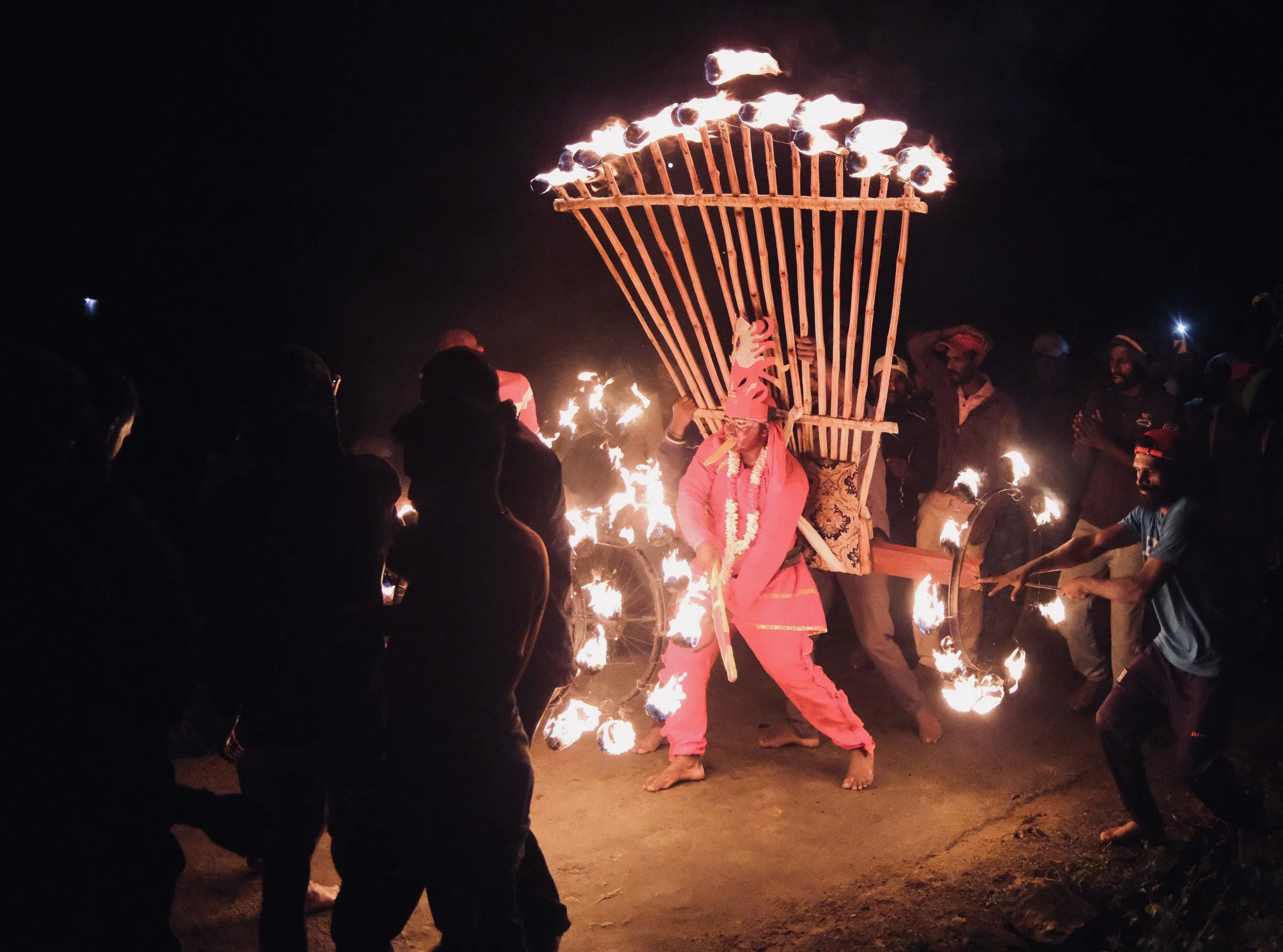 A nighttime scene of a traditional fire dance, with a performer dressed in pink and a large, flaming wooden structure shaped like a basket or crown, surrounded by a crowd of spectators and other participants holding torches or fire props.