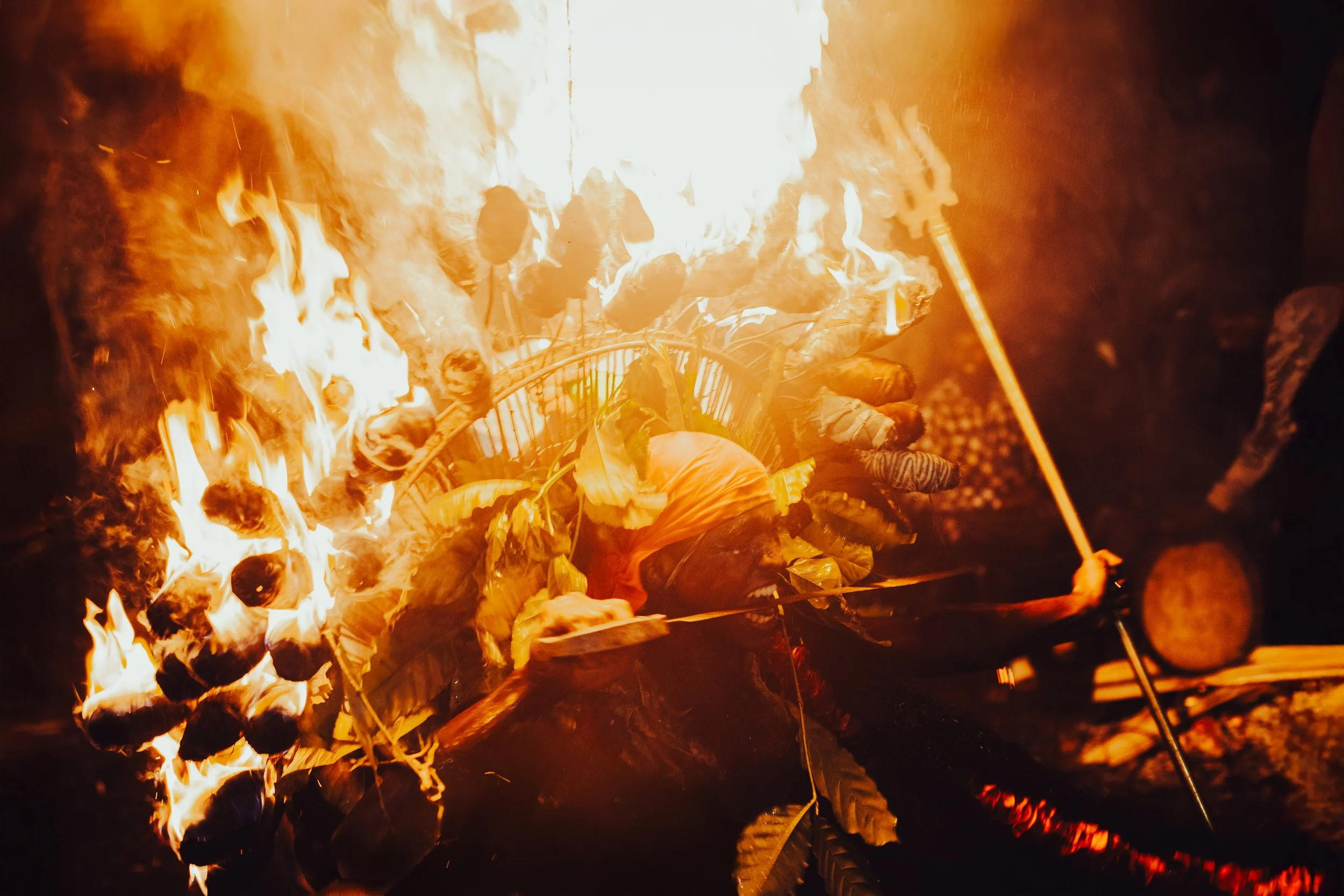 A person wearing traditional indigenous clothing and a large feathered headdress is performing a fire dance, holding a lit torch in front of a large fire with flames and smoke.