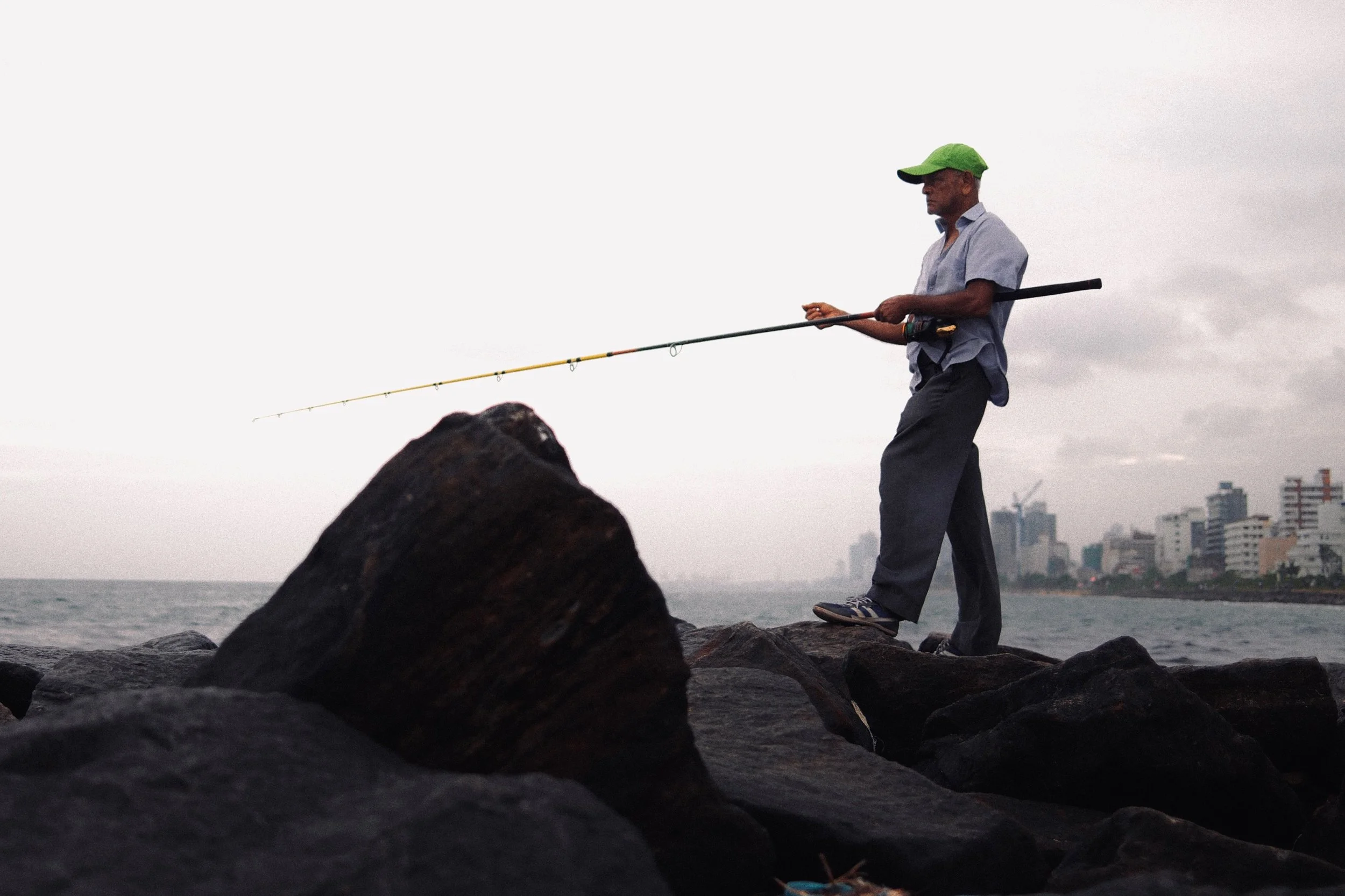 Man fishing on rocks by the ocean with city buildings in the background during overcast weather.