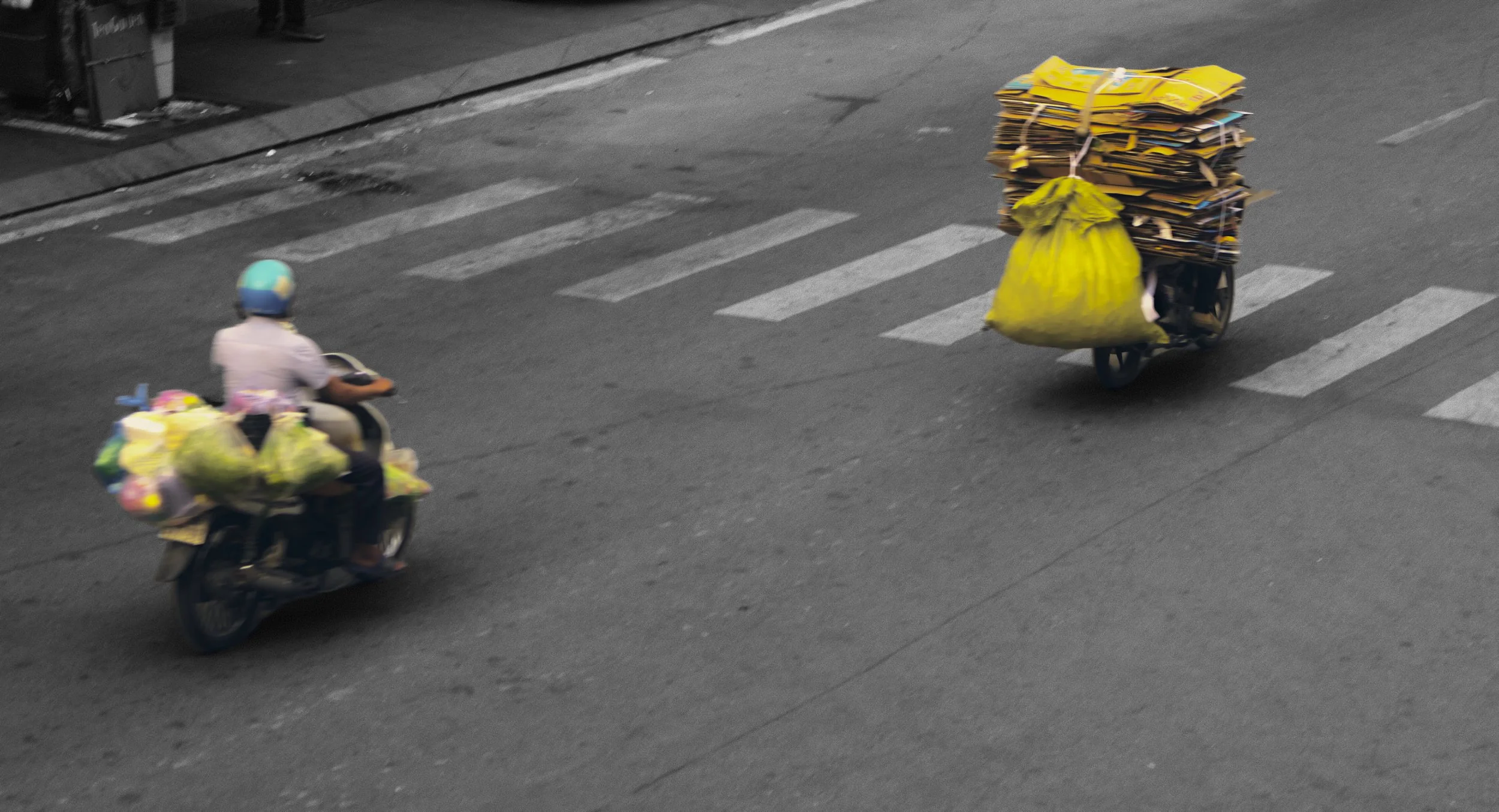 Two motorbikes riding on a city street with large piles of boxes and yellow bags attached to their backs, crossing a zebra pedestrian crosswalk.