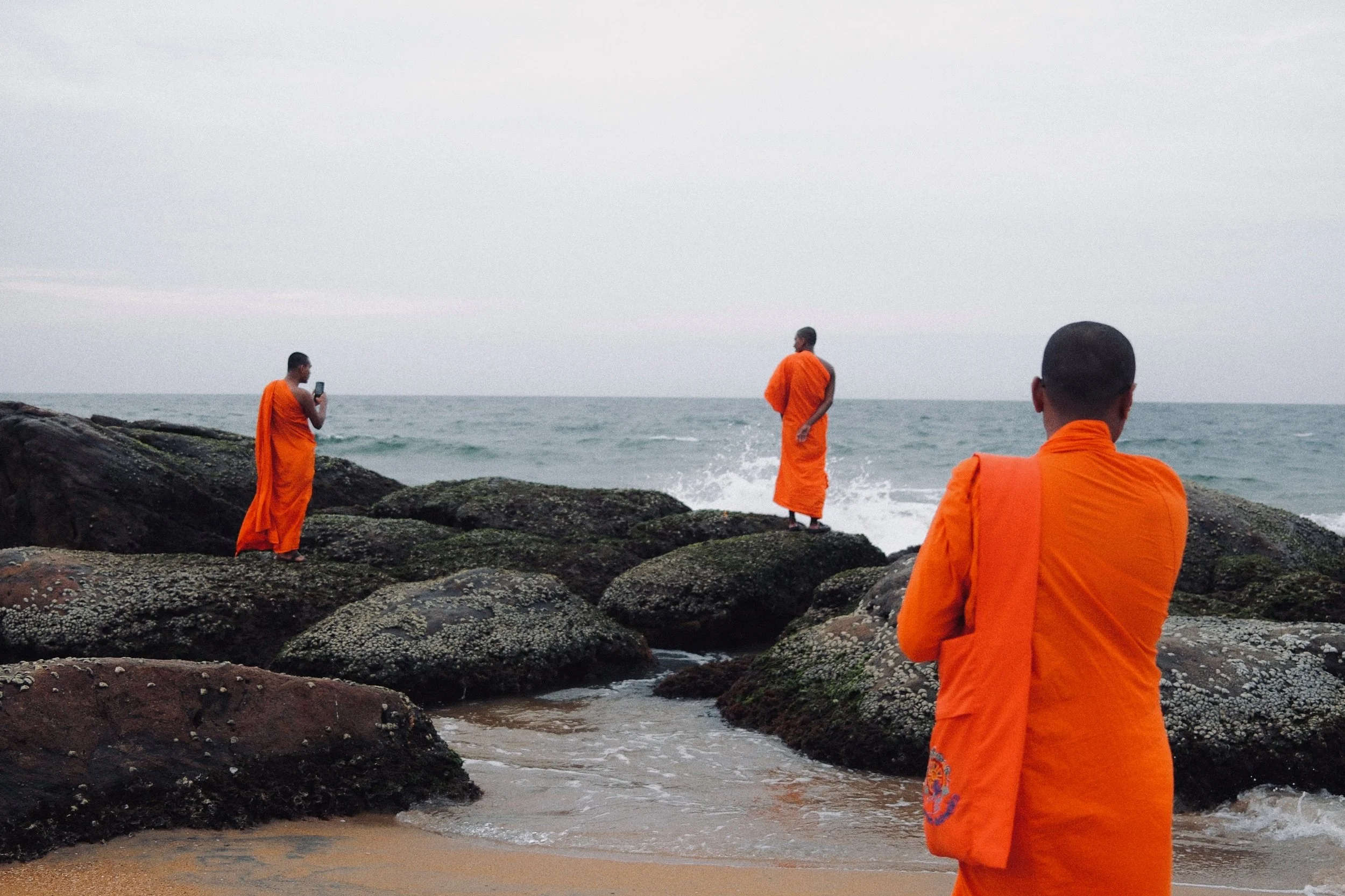 Three monks dressed in orange robes on rocks at the beach, with the ocean and a cloudy sky in the background.