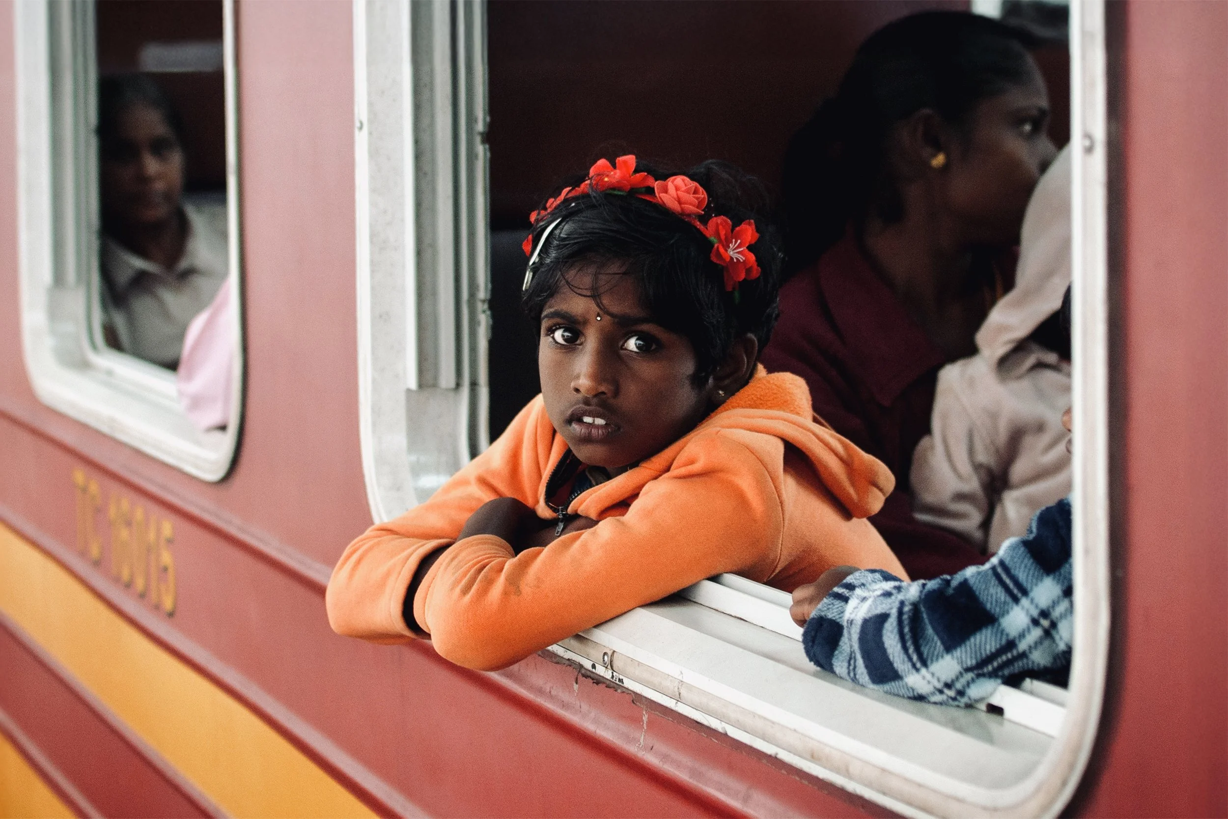 A young girl with a red floral headband and orange hoodie leaning out of a train window, looking at the camera with a curious expression, with other children visible inside the train.