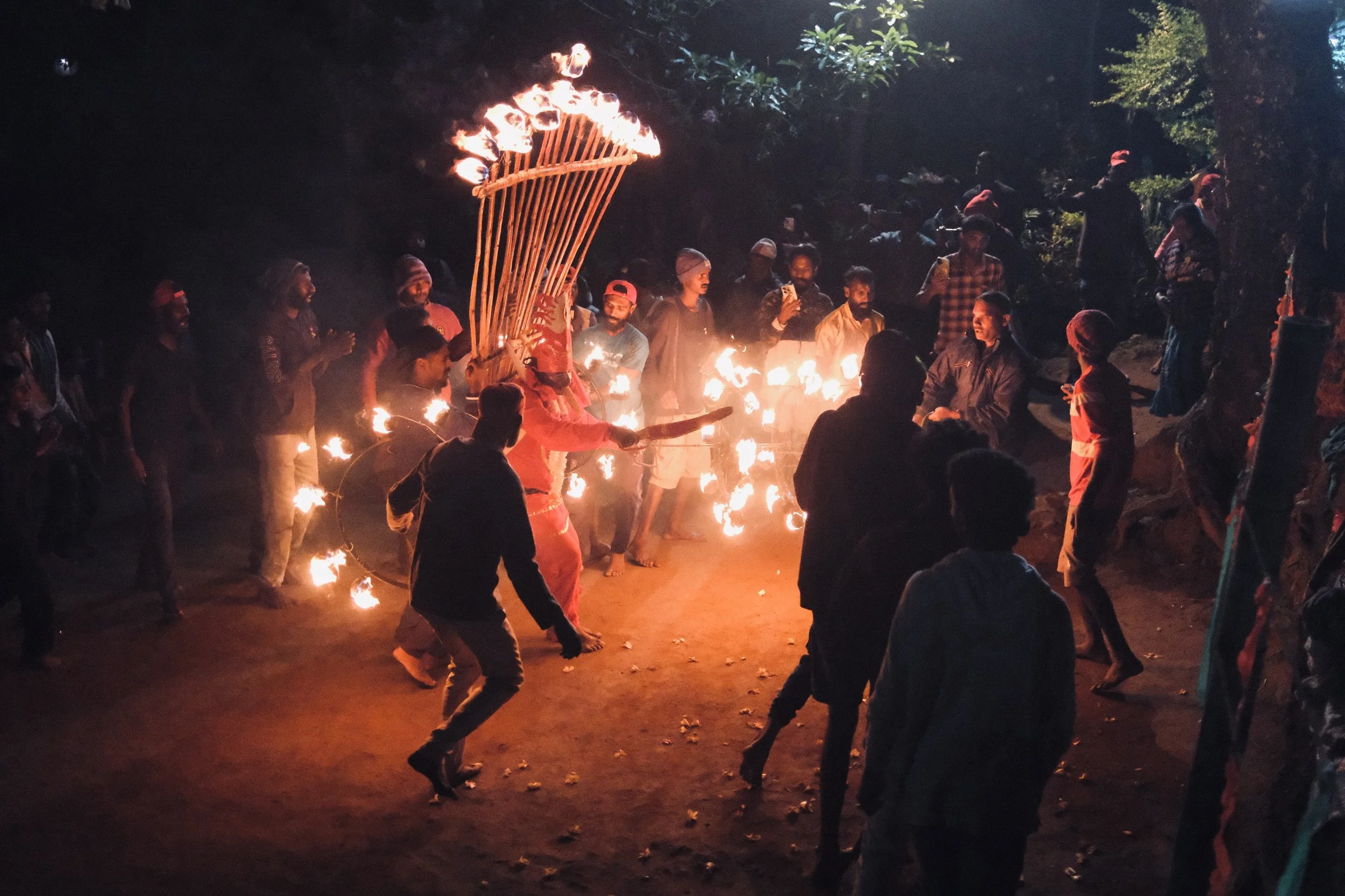 People gathered around a fire performer creating fire during a night-time celebration. The performer has fire torches and is surrounded by onlookers in an outdoor setting with trees.