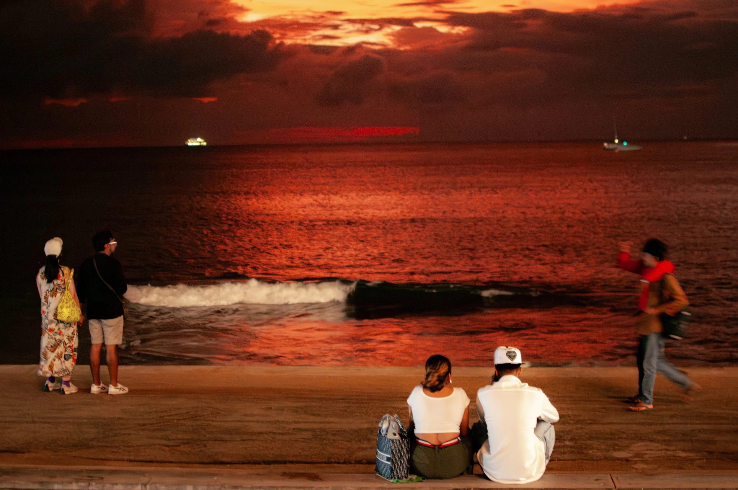 People on a beach during a vibrant red and orange sunset, looking out at the ocean with boats in the distance.