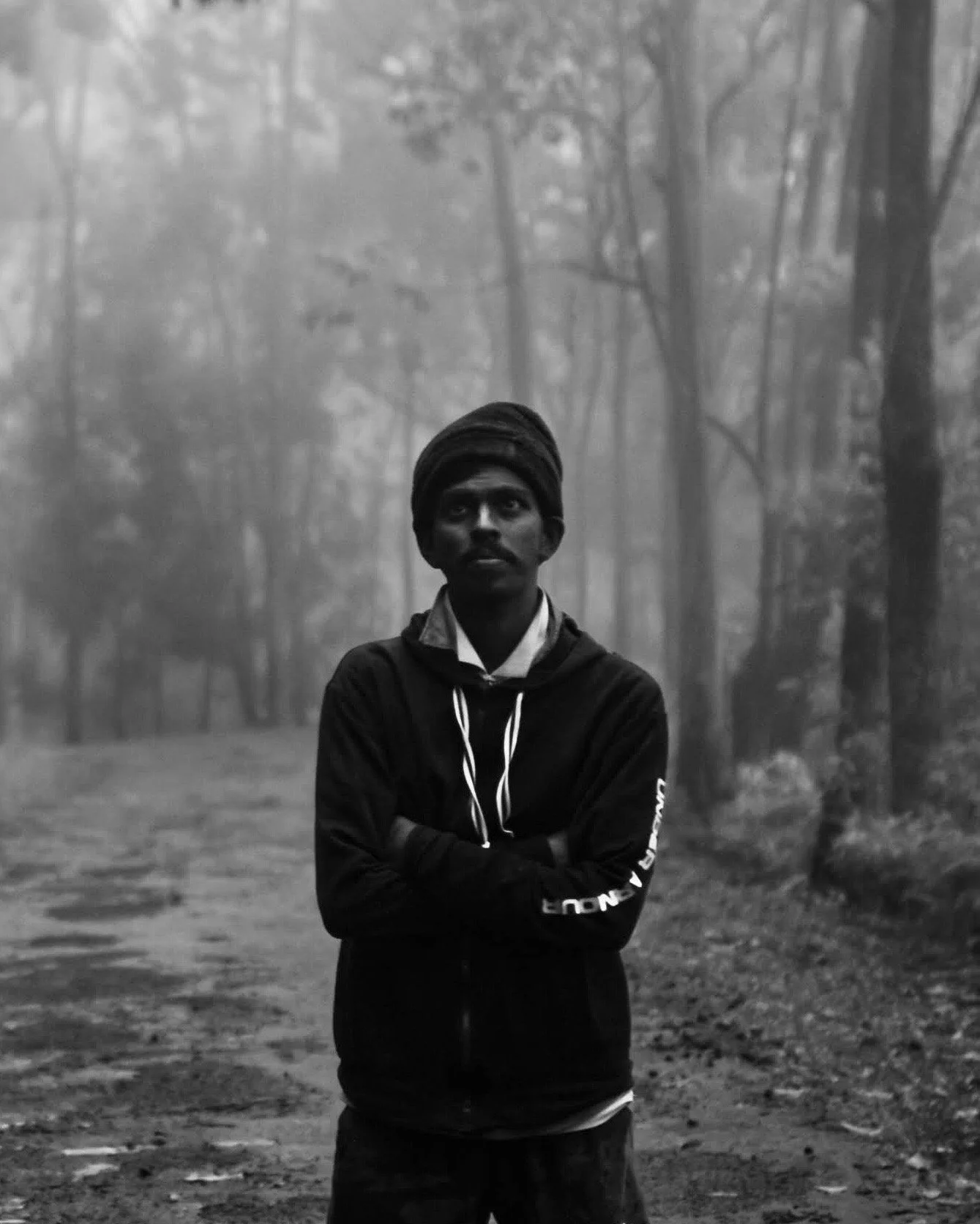 A young man standing with arms crossed in a foggy forest, wearing a head wrap and a hoodie.