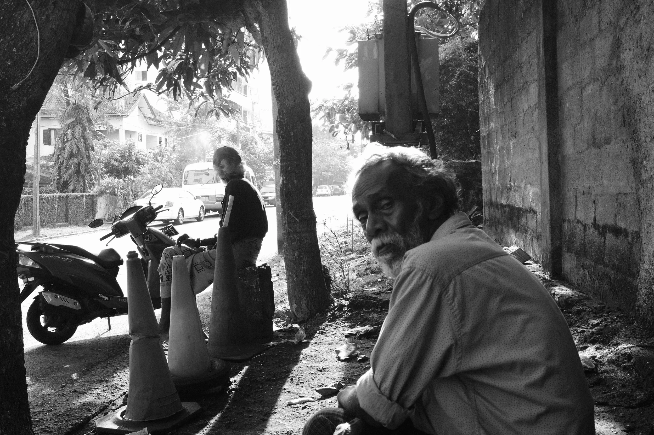 An elderly man with a beard sitting on the ground near a brick wall, looking at the camera, with a woman sitting on a concrete block near parked bikes and traffic cones, in an urban street scene. Bright sunlight creates silhouettes and shadows.