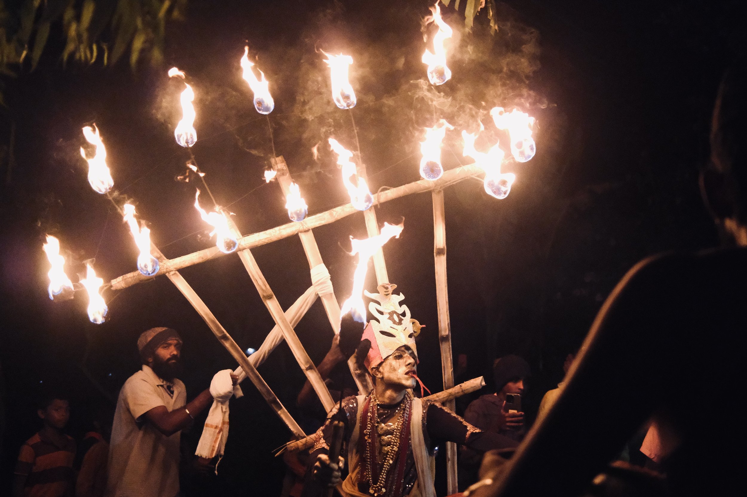 People participating in a nighttime cultural or religious festival showing a performer with face paint and a costume, holding a large wooden structure with multiple flames on it, surrounded by onlookers.