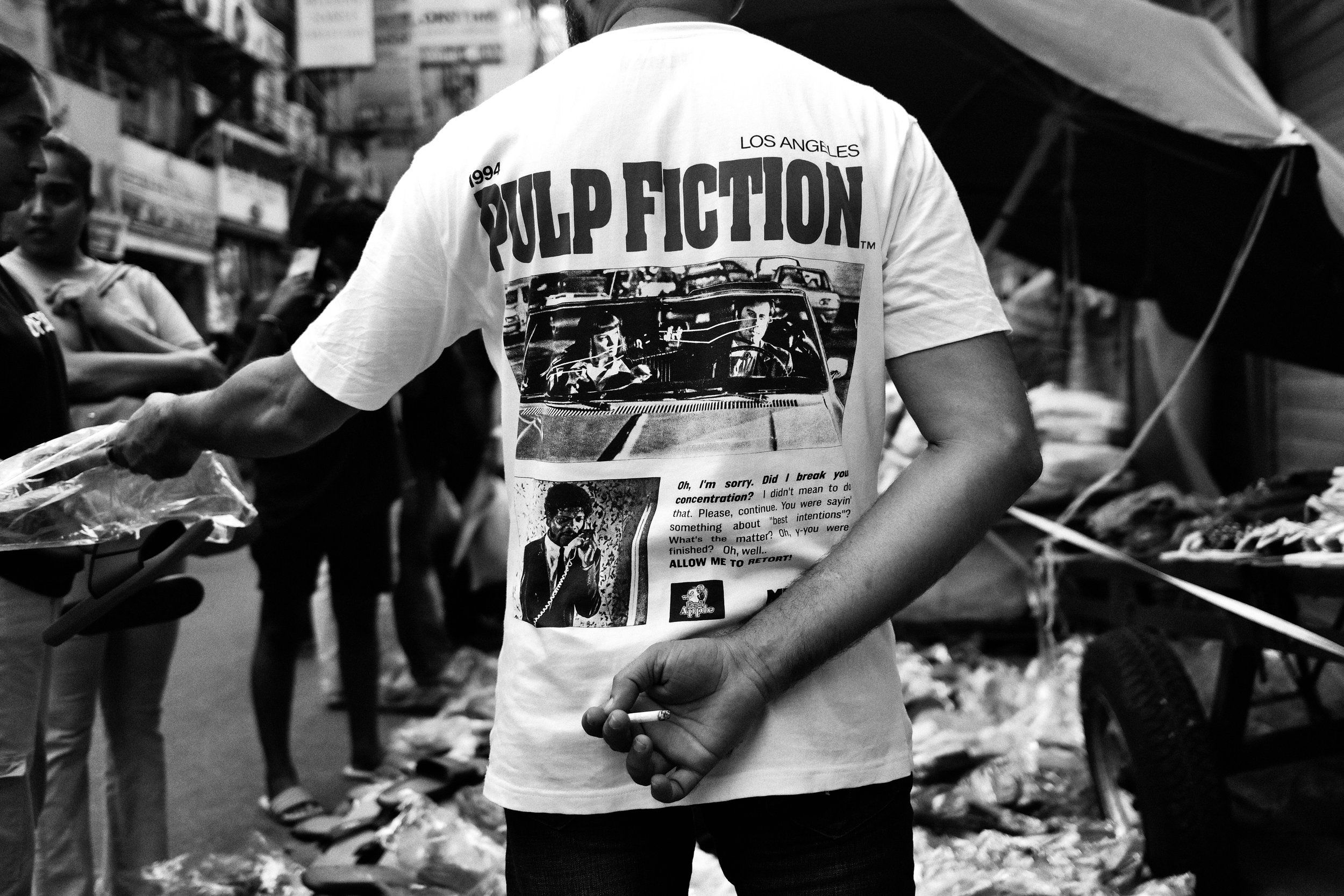 A man wearing a white T-shirt with the 'Pulp Fiction' movie poster in black and white, standing in a busy street market. The image captures his back, with other people and market stalls visible in the background.