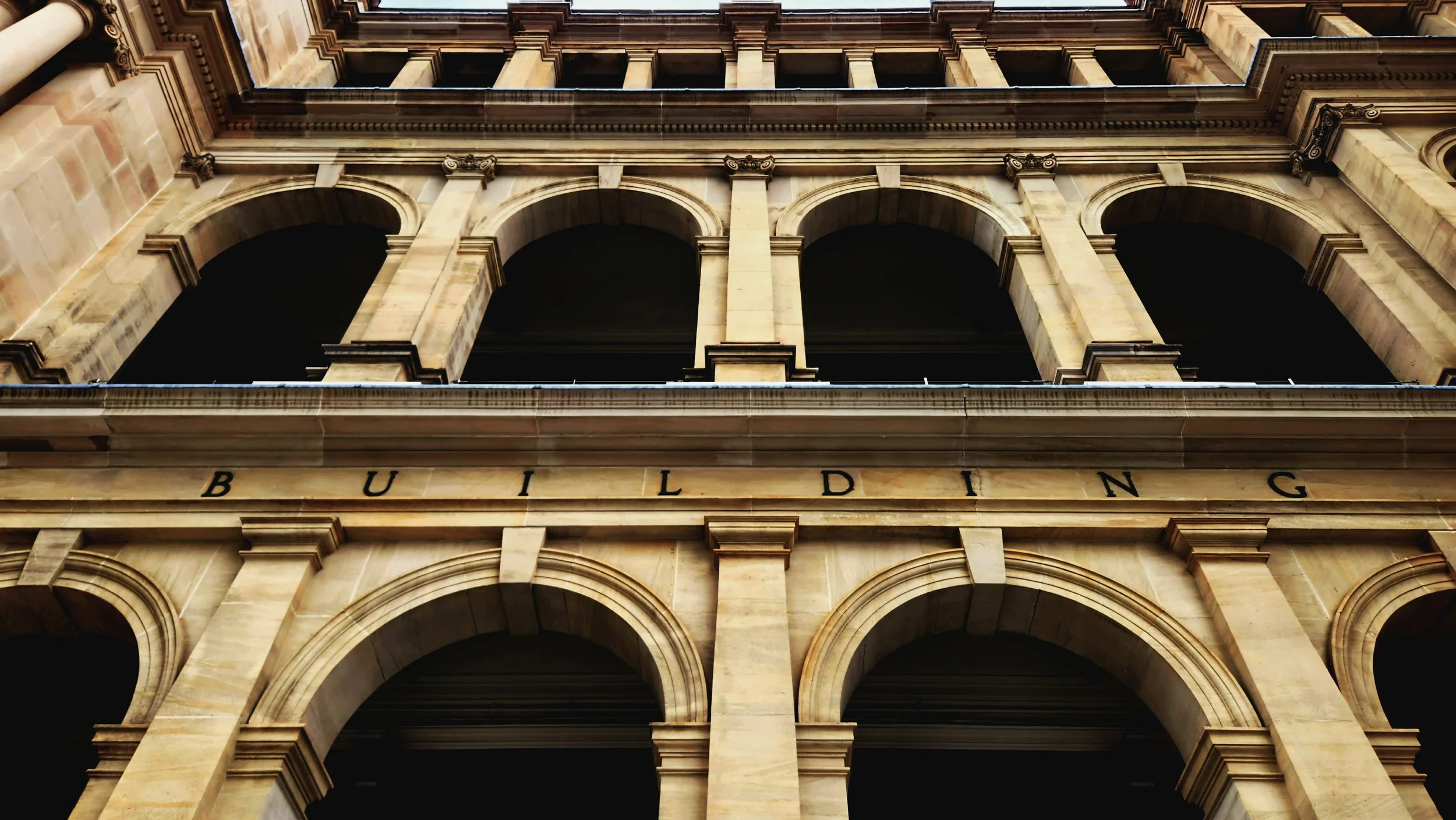 Close-up view of a historic building's stone facade with arched windows and detailed columns, and the word "BUILDING" engraved on the stone below the arches.