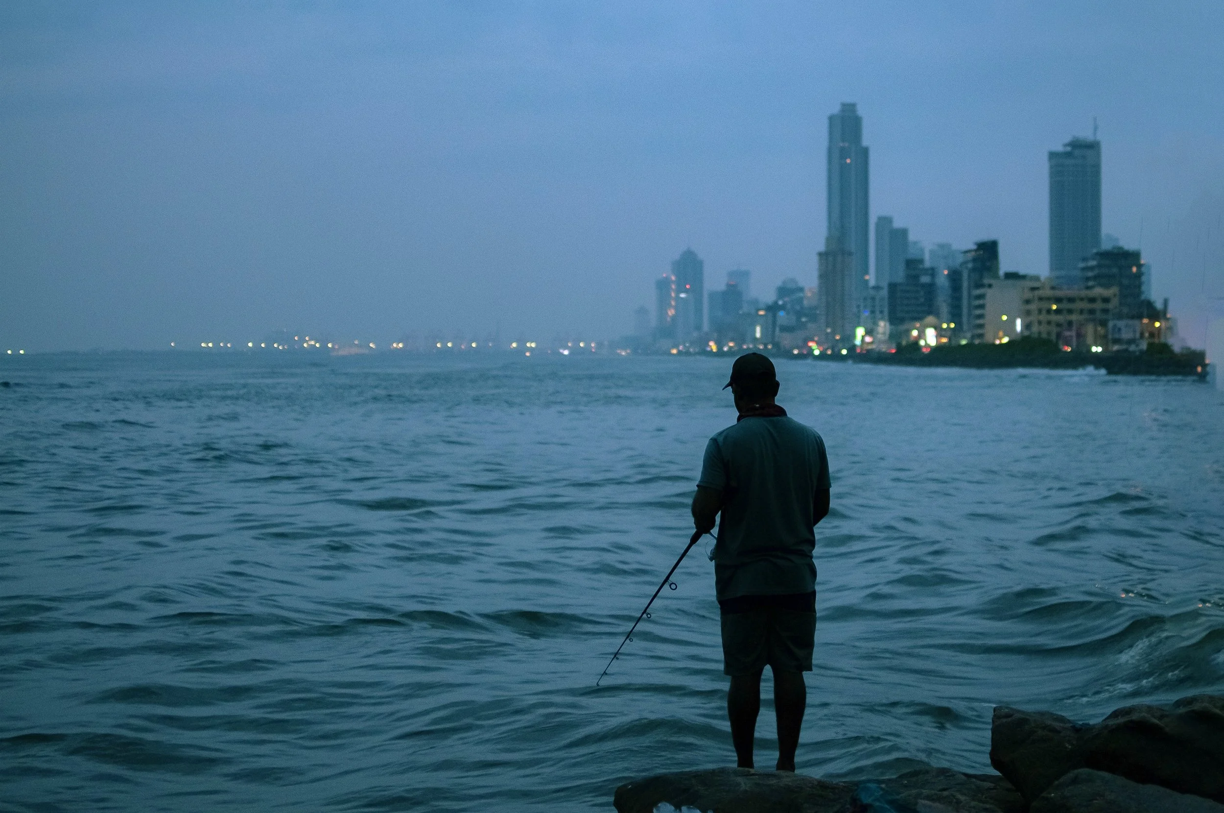A man fishing on rocks by the water at dusk, with a city skyline featuring tall buildings illuminated in the background.