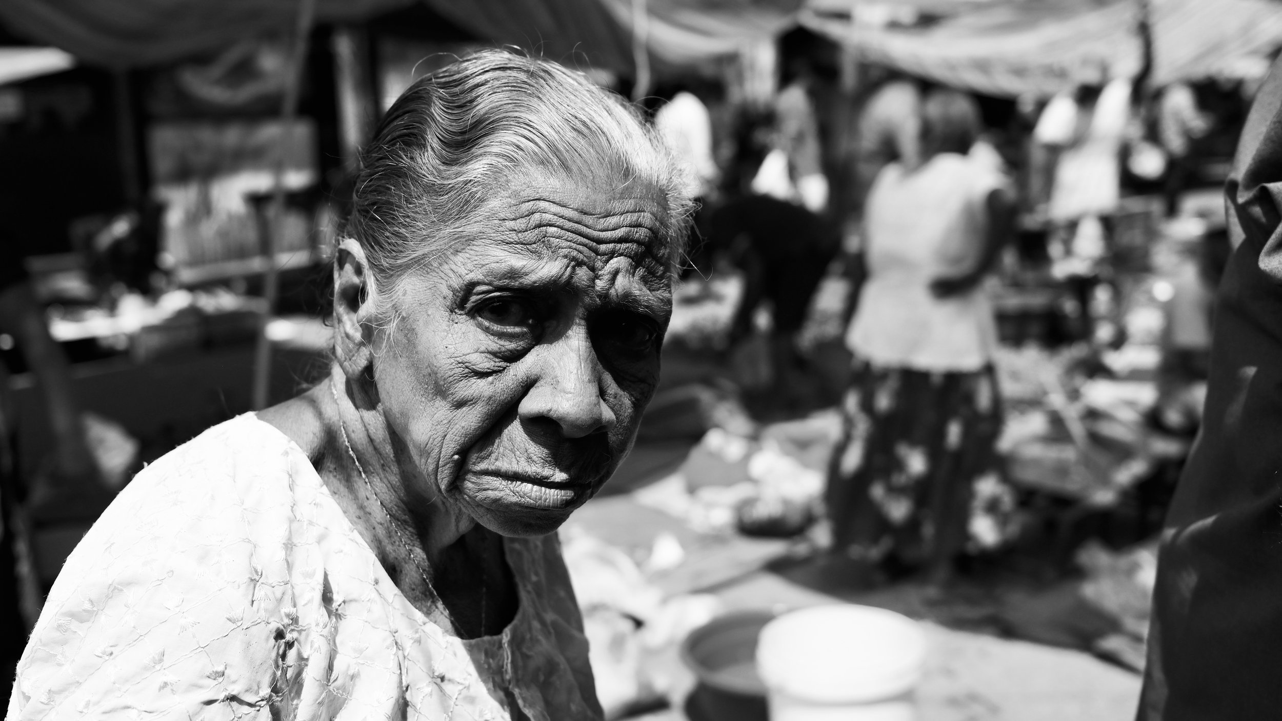 An elderly woman with wrinkled skin and gray hair, looking directly at the camera in a marketplace with blurred figures and market stalls in the background.