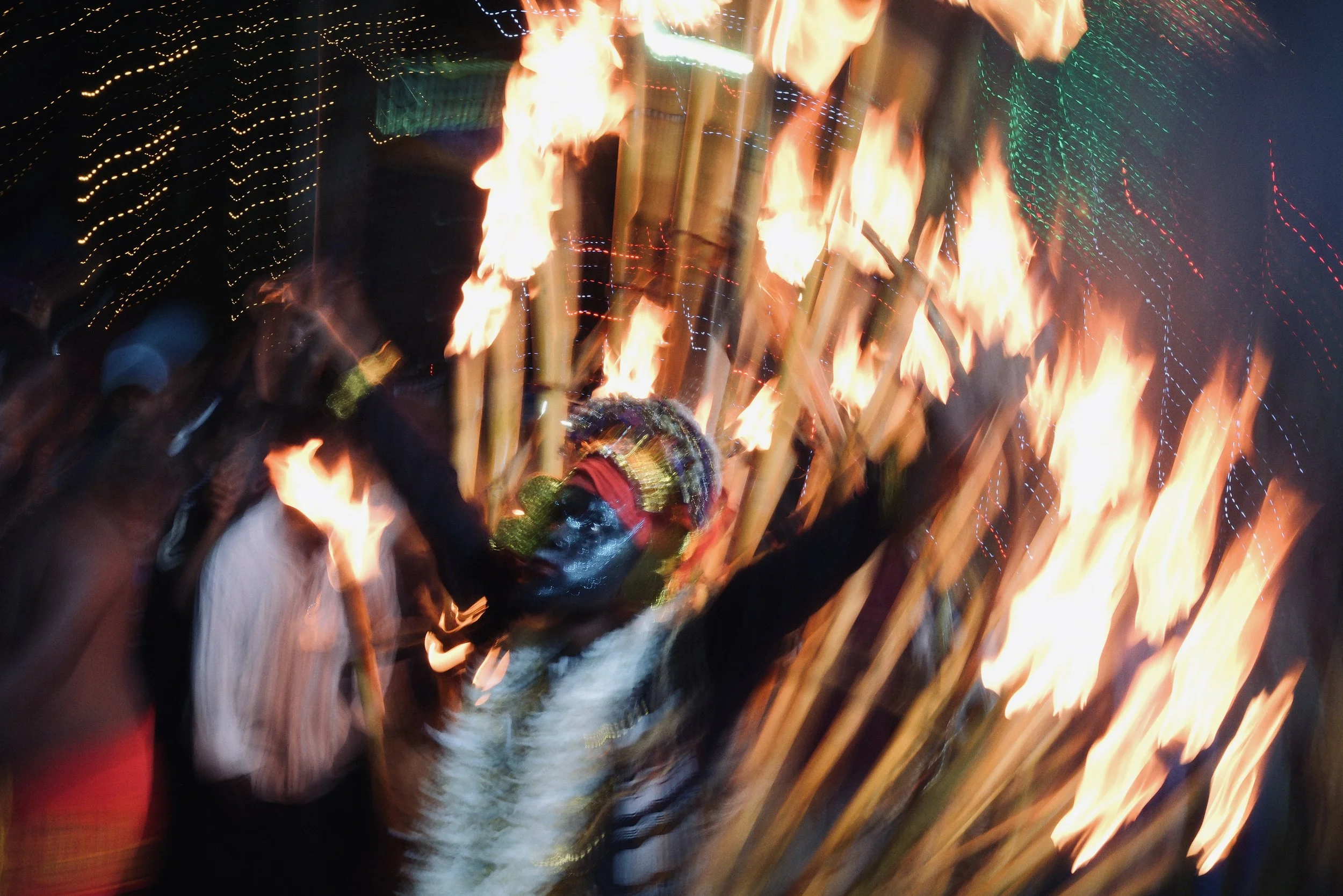 A person dressed in traditional attire with face paint, holding torches, participating in a nighttime cultural celebration with colorful lights and blurred motion.