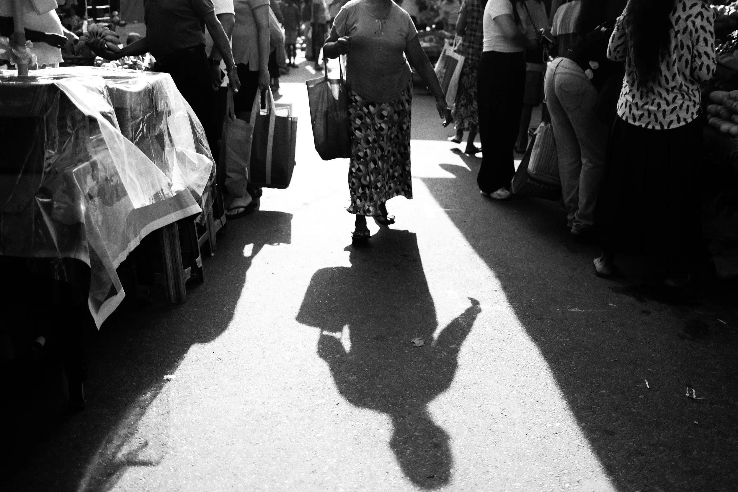 A person walking through a bustling outdoor market with vendors and shoppers, casting a shadow on the ground.