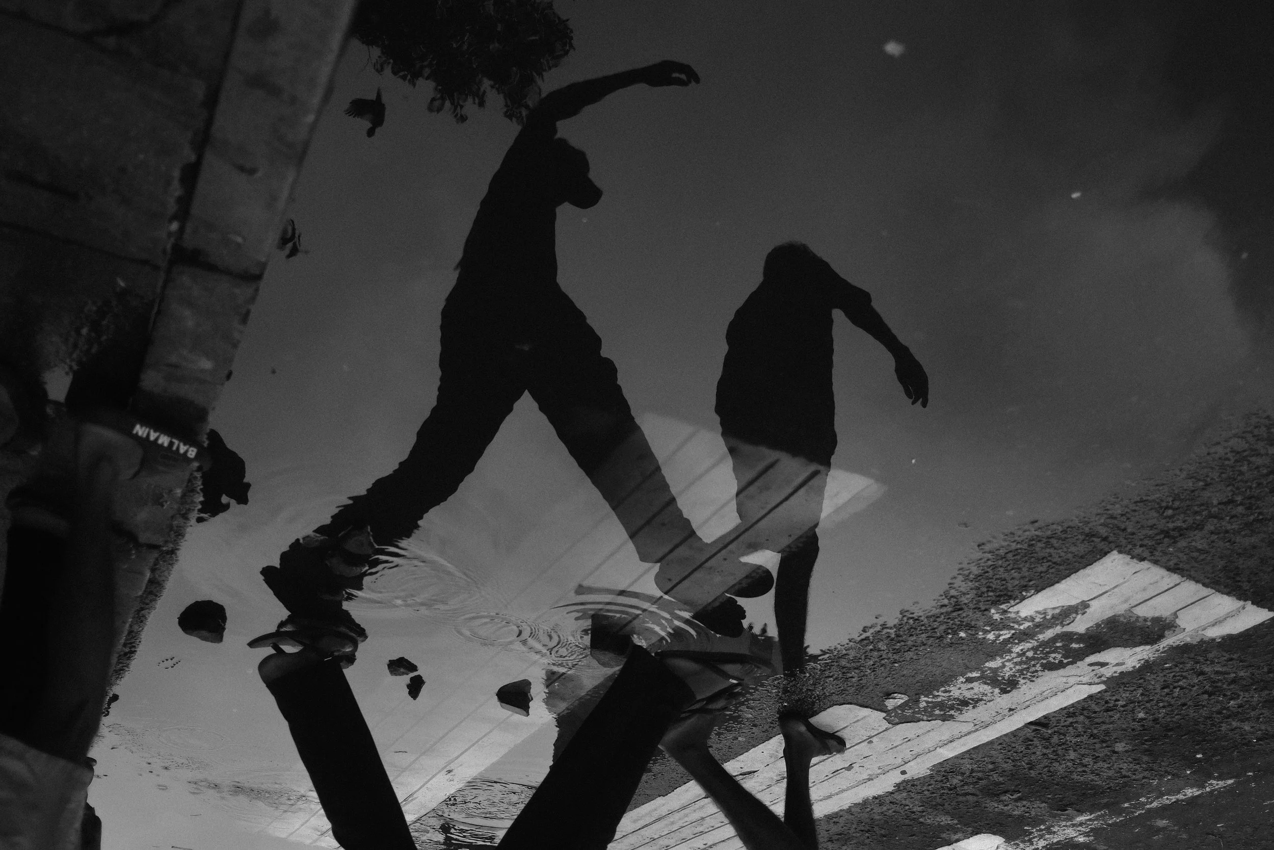 Reflection of two people, possibly skiers or snowboarders, in a puddle or water on the ground, surrounded by snow and winter environment.