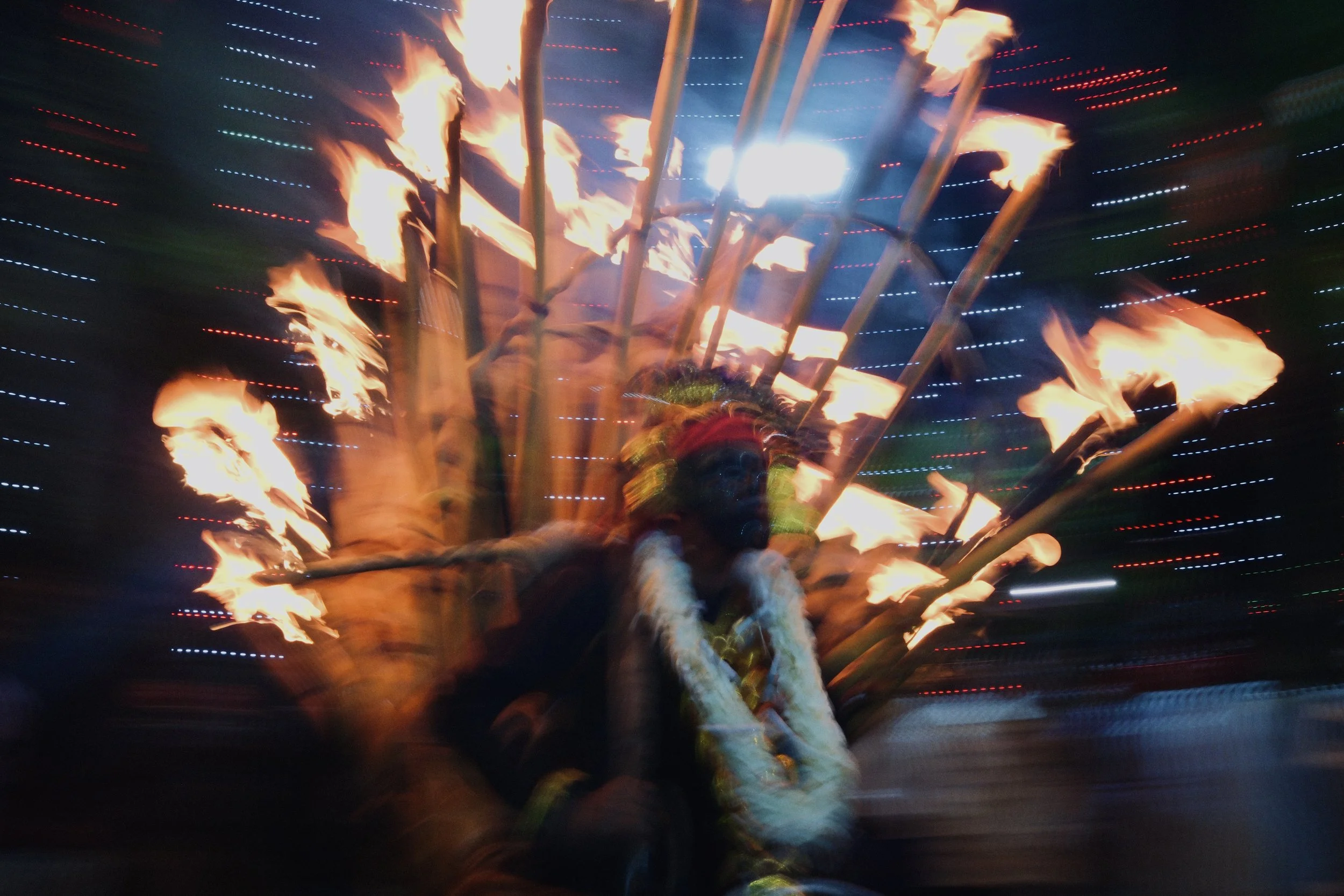 A person dressed in traditional attire with a headdress and floral necklace, participating in a cultural or religious festival involving fire spinning or flaming performance at night with colorful lights in the background.