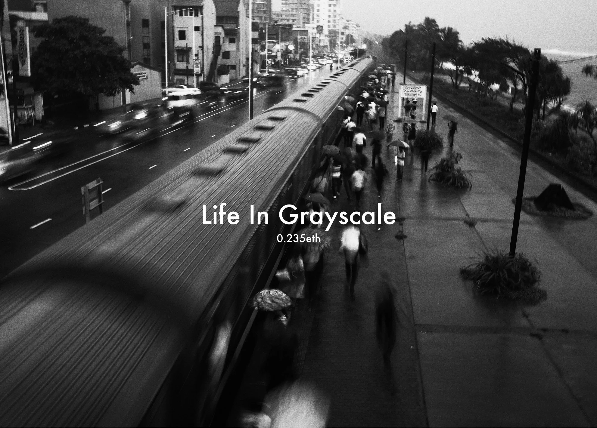 A grayscale photo of a busy city street. A train is in motion on the left side, with people walking on the sidewalk on the right side, some holding umbrellas. Tall buildings line the street, and trees are visible along the sidewalk.