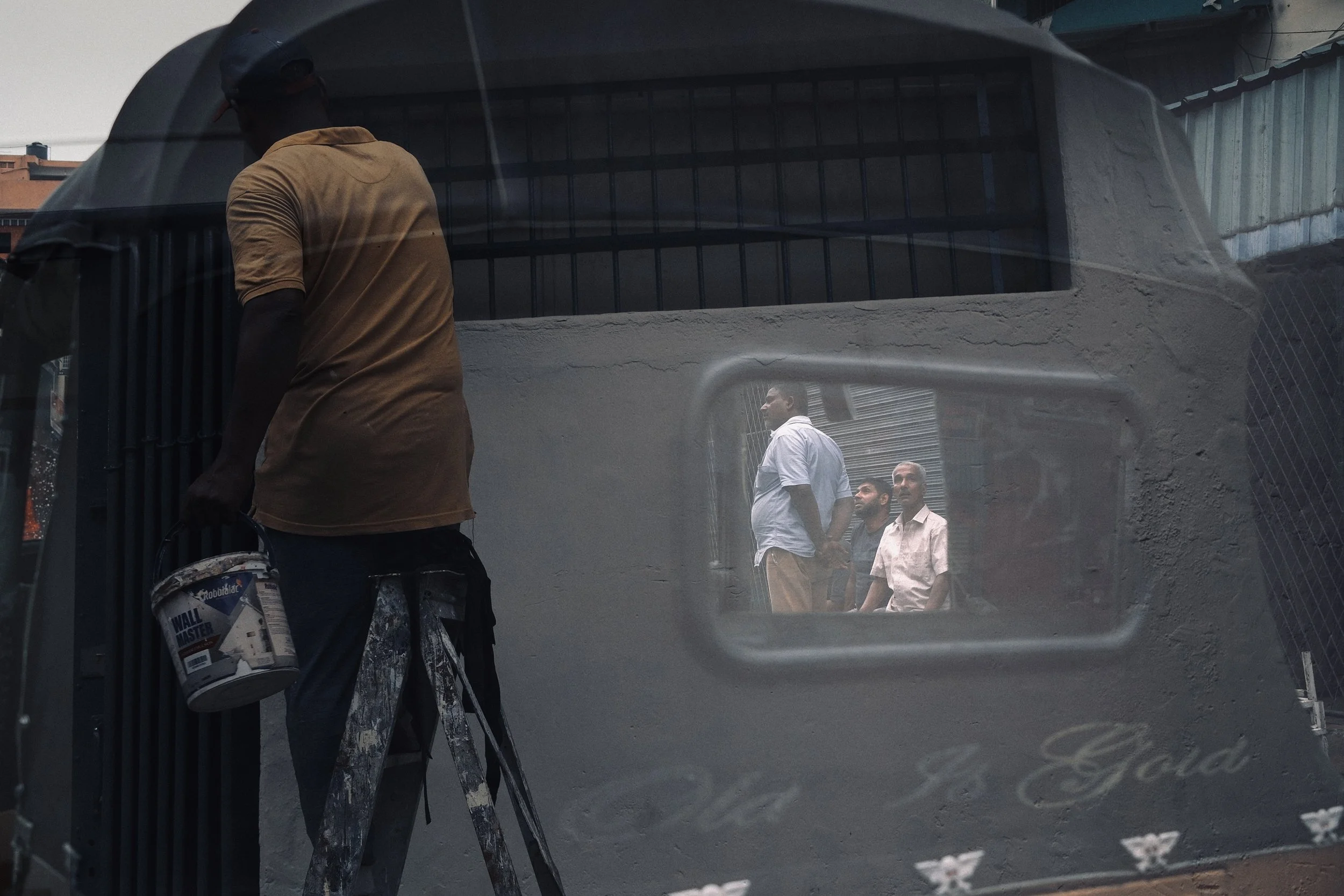 Reflected image of three men standing in line and looking in a direction, seen through a window of a building with a rough wall, alongside a man on a ladder holding a bucket of paint.