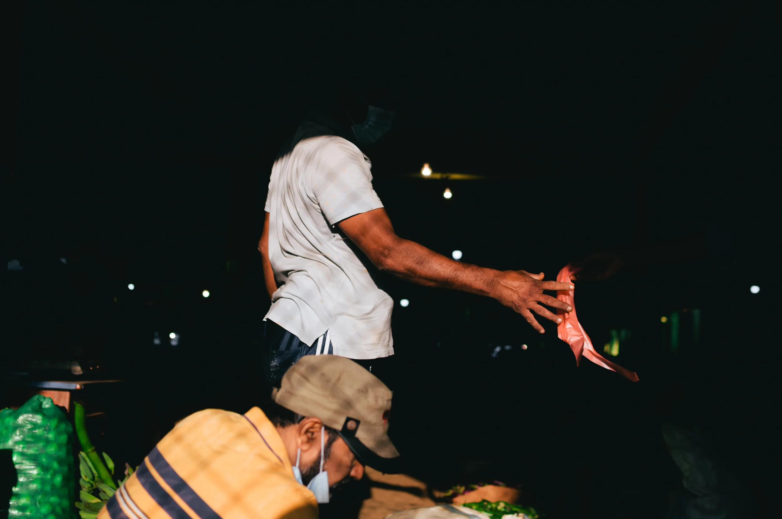 Man in a white shirt and face mask with dark hair stands at night, holding a pink plastic bag, while another man in a tan cap and striped shirt wears a mask and analyze items on a table, with a dark background and streetlights.