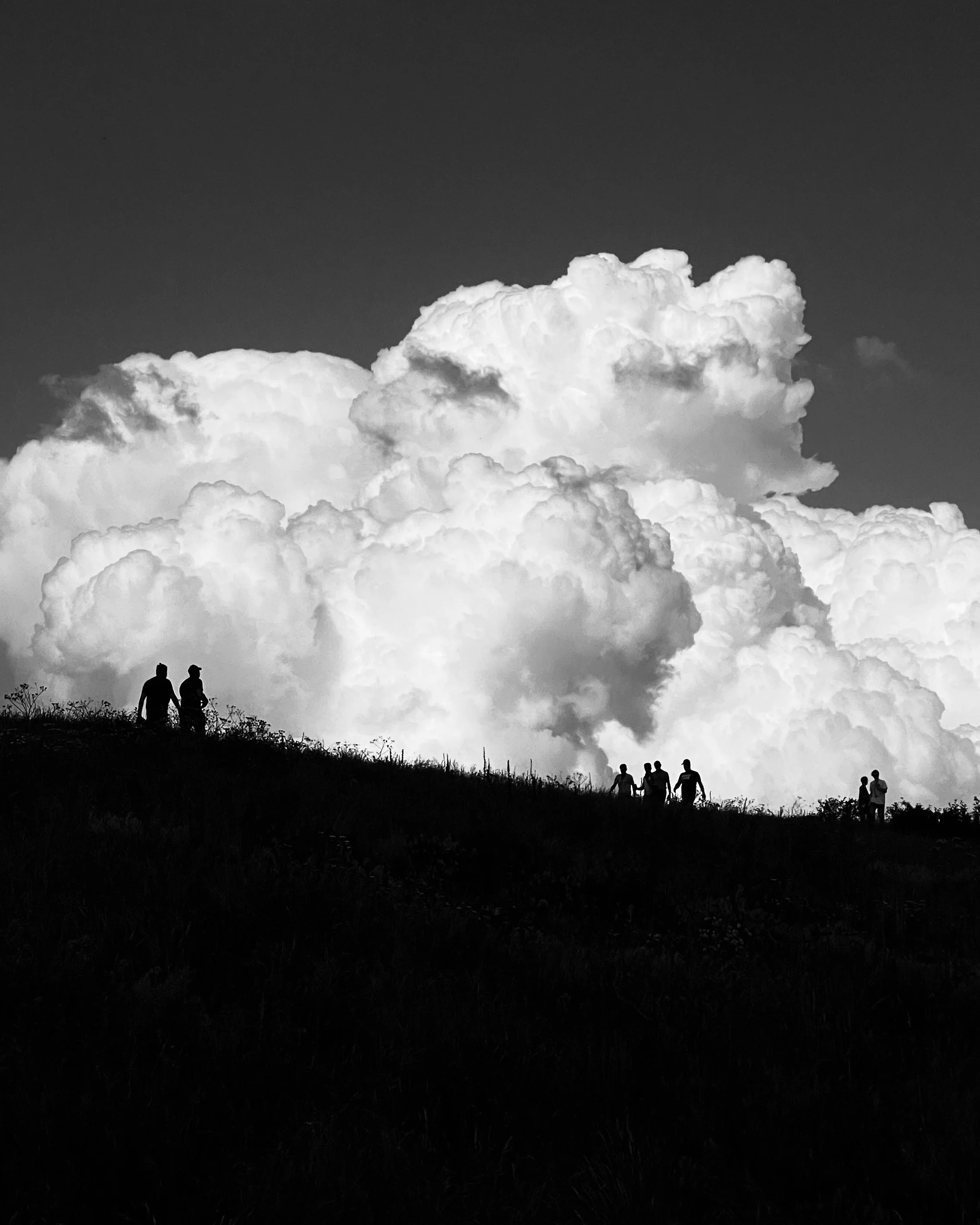 Black and white photograph of a group of people sitting and standing on a hilltop with a dramatic cloud-filled sky in the background.