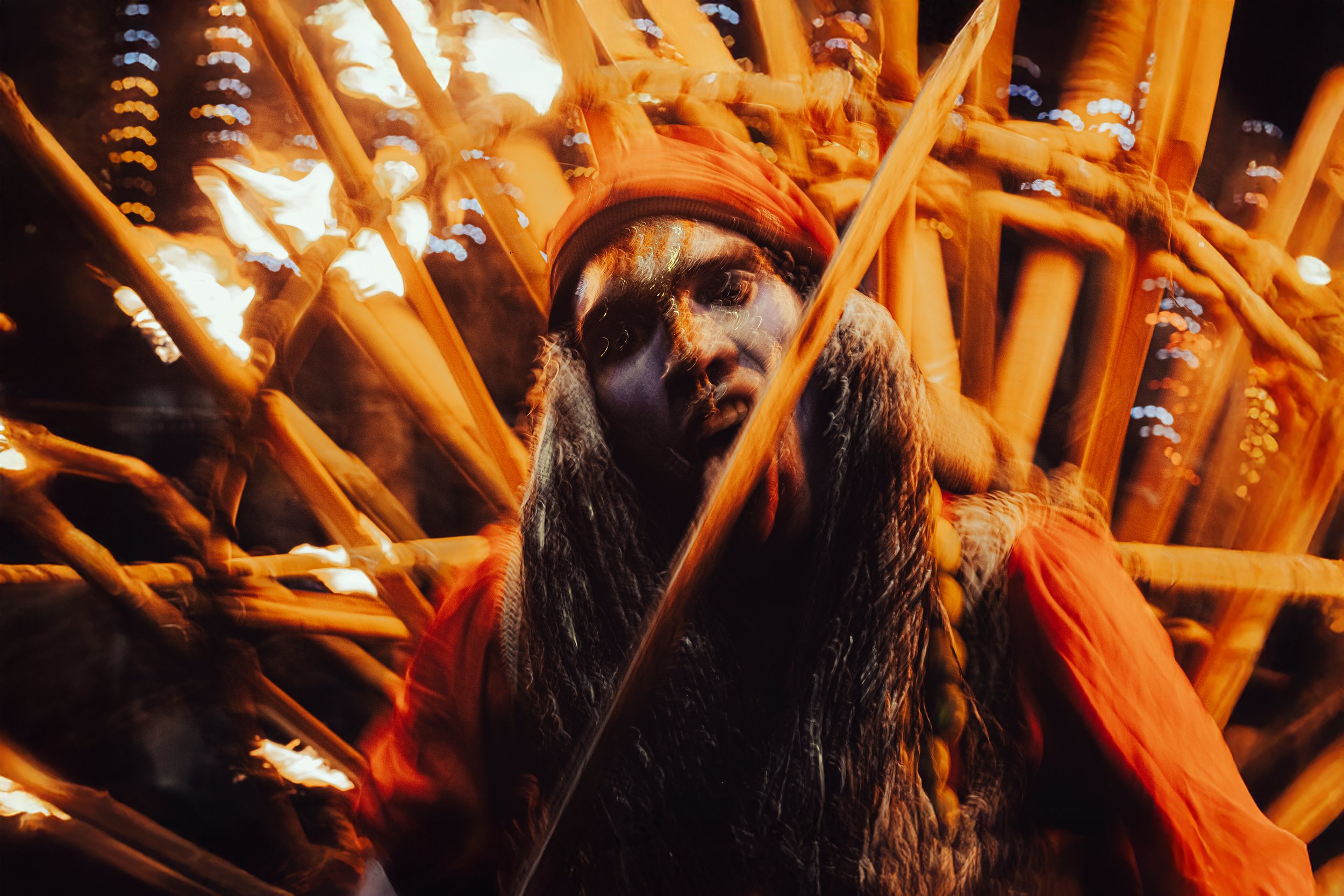 A person wearing an orange jacket and beanie, with long hair and face paint, is surrounded by flames and wooden sticks at night, possibly part of a fire performance or ritual.