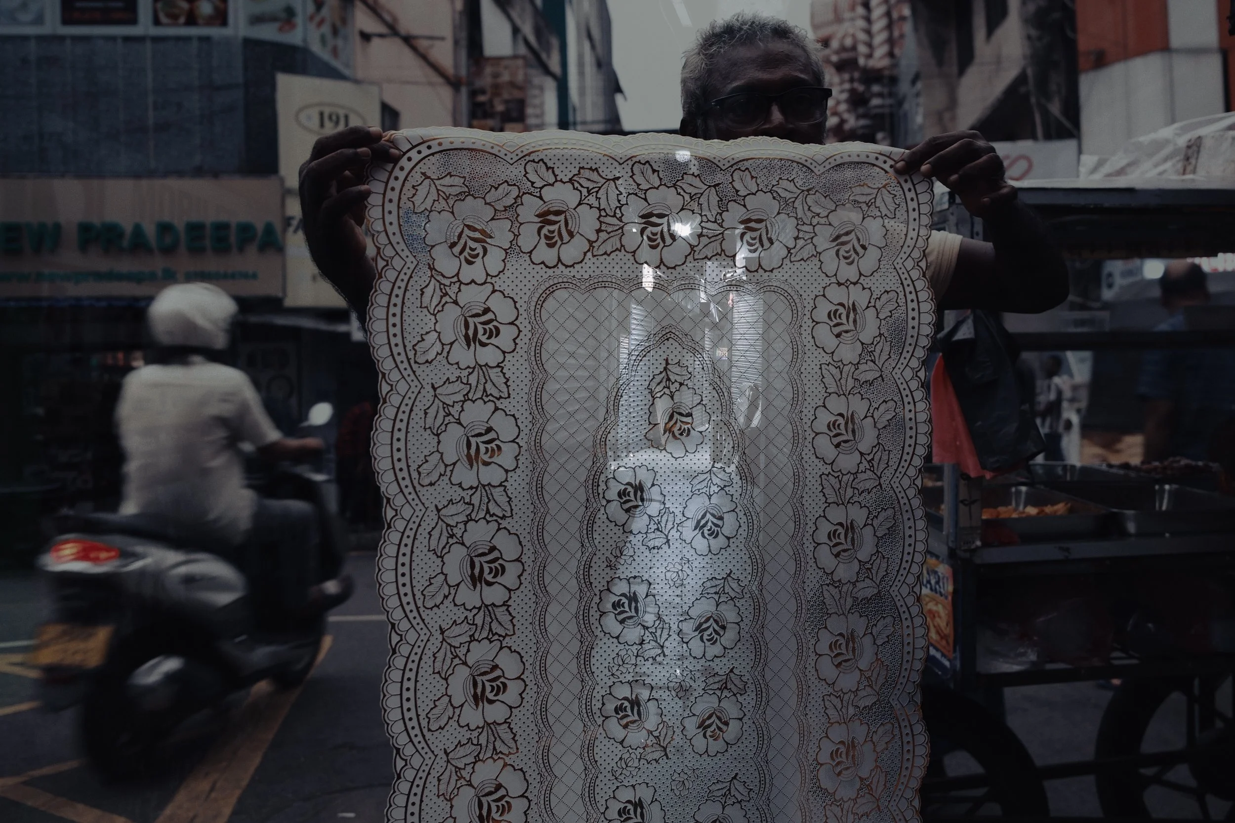 A person holds a large ornate lace fabric with floral patterns, with the street scene and a motorcycle in the background.