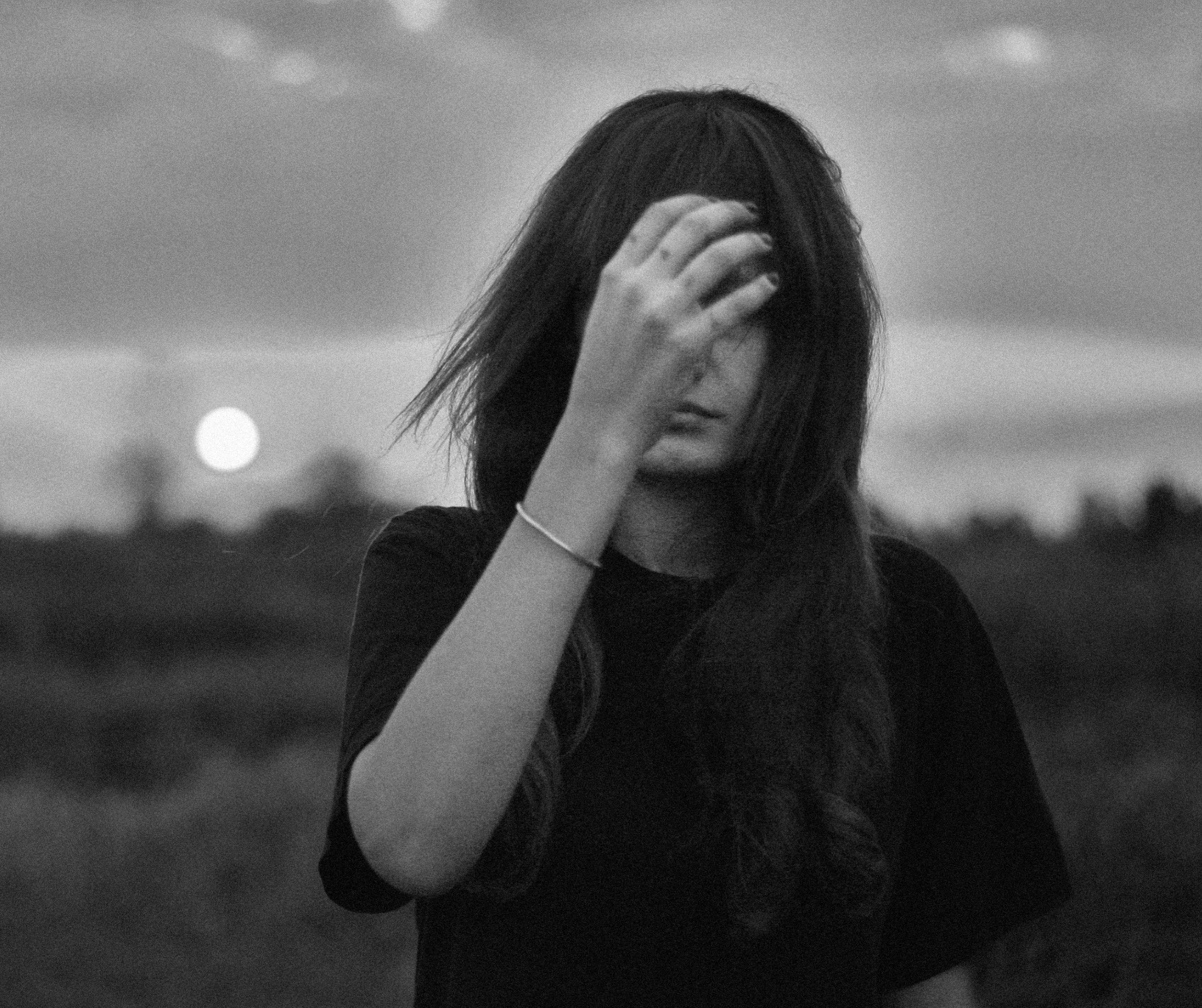 A woman with long hair covering her face, holding her hand to her forehead, outdoors at dusk or dawn, black and white photo