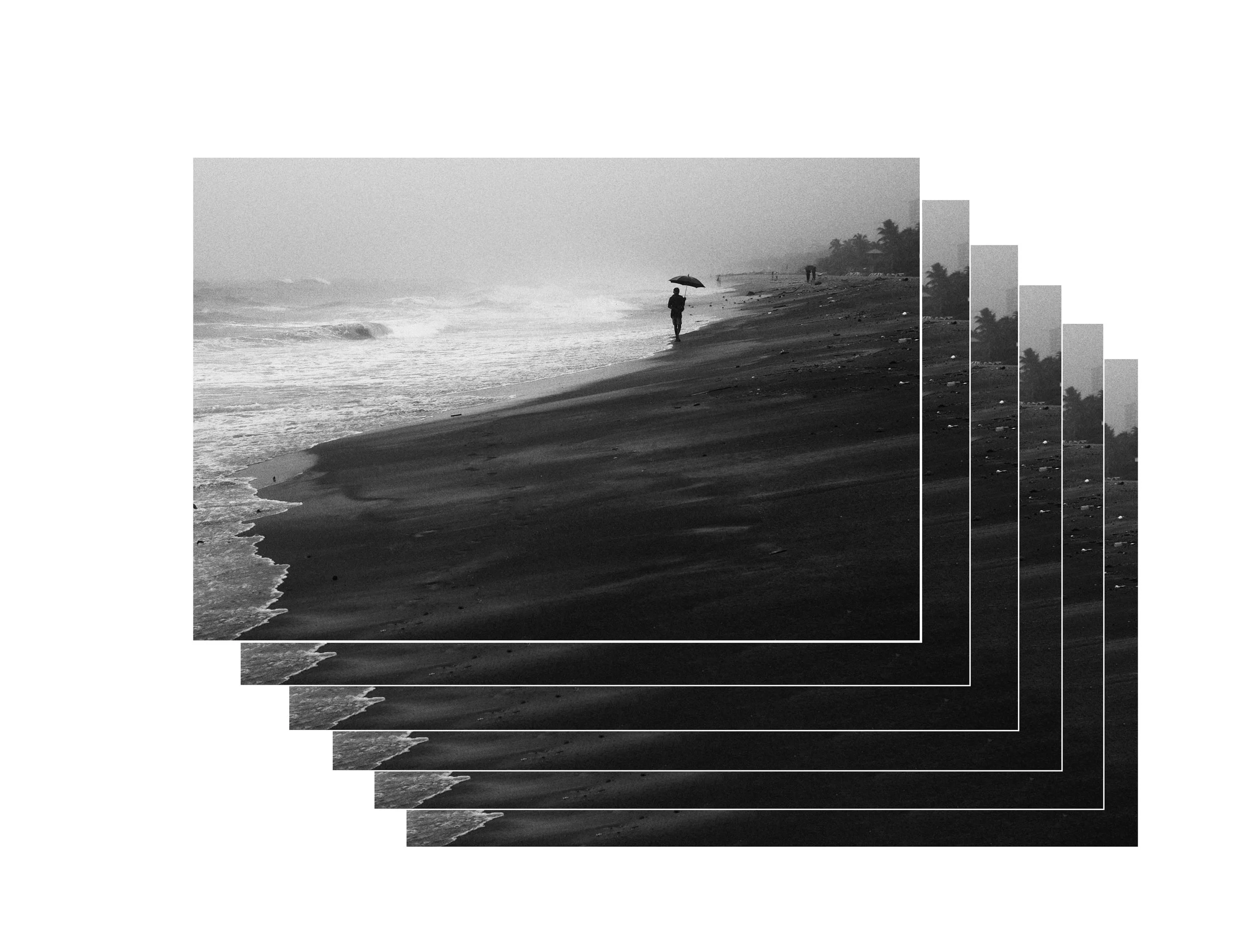 A person walking along a beach holding an umbrella, with trees and a few other people in the distance, in black and white.