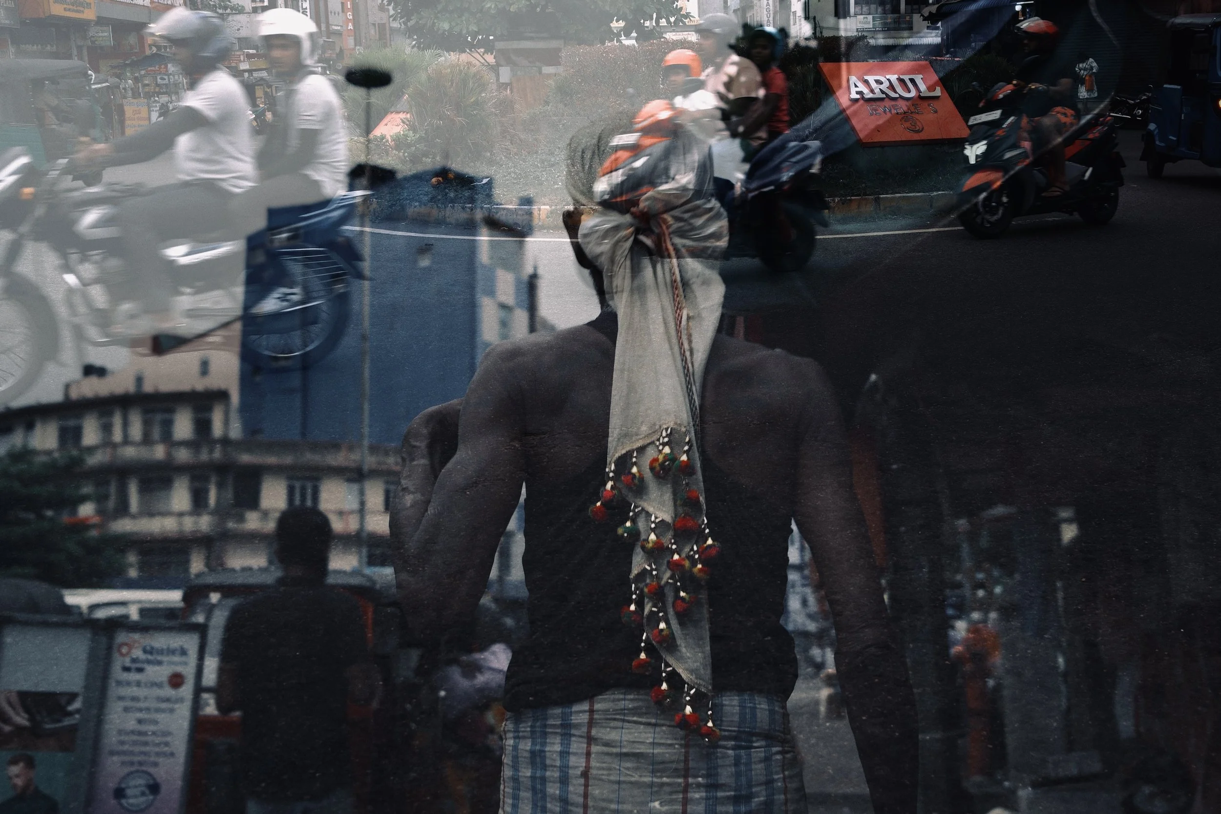 A reflection of a man with a headscarf and jewelry on her back, seen through a glass window, with a busy street scene of motorbikes and people in the background.