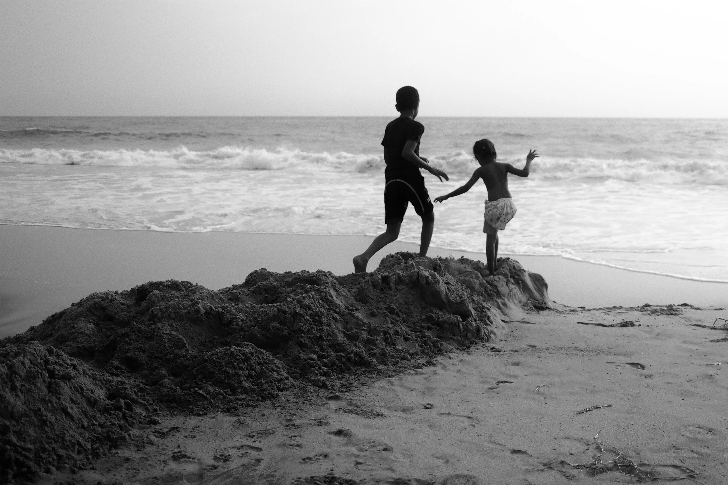 Two children, a boy and a girl, playing on a mound of sand at the beach near the water, with ocean waves in the background, in black and white.