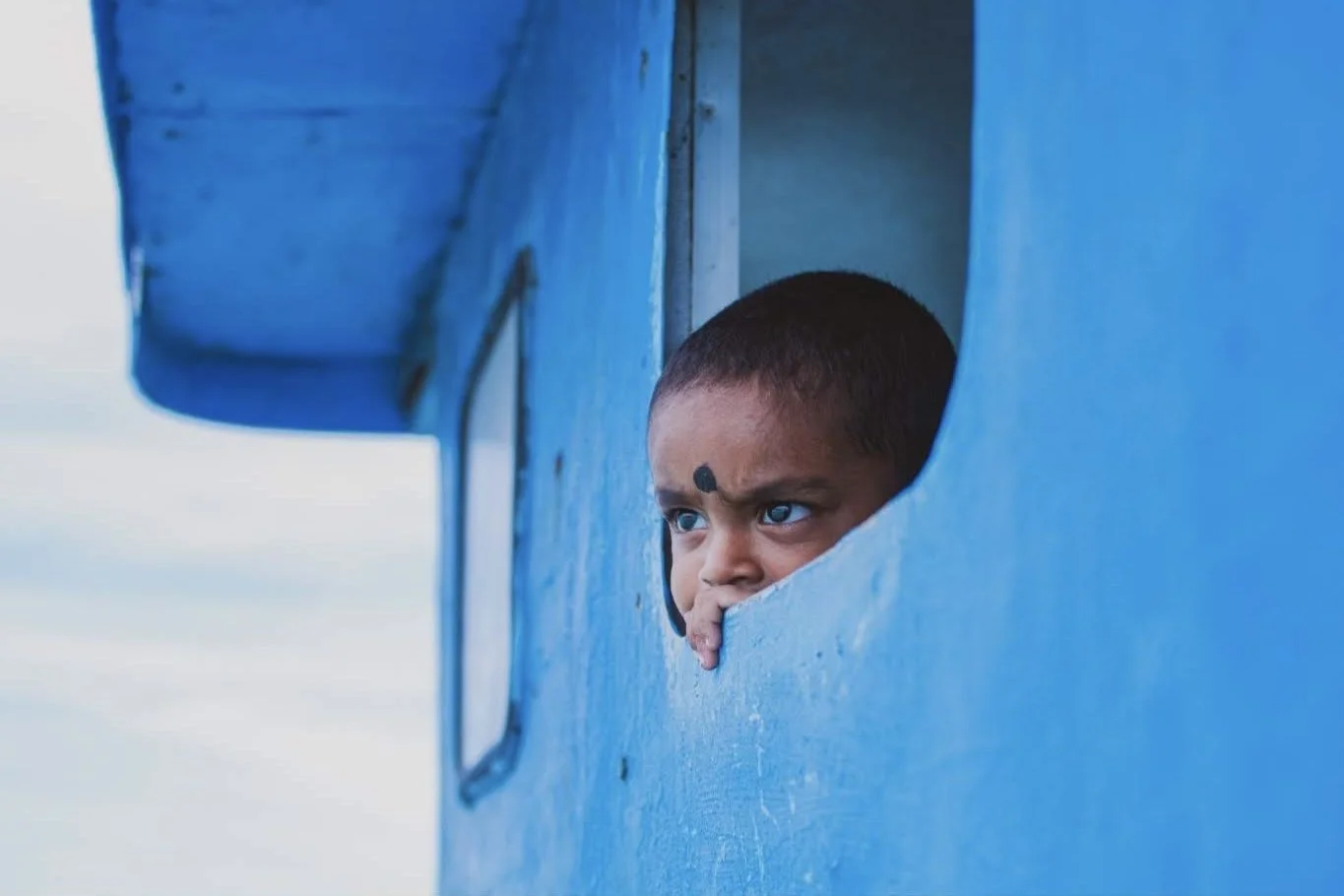 A young boy with short hair and a black bindi on his forehead looks out from a small window in a blue structure.