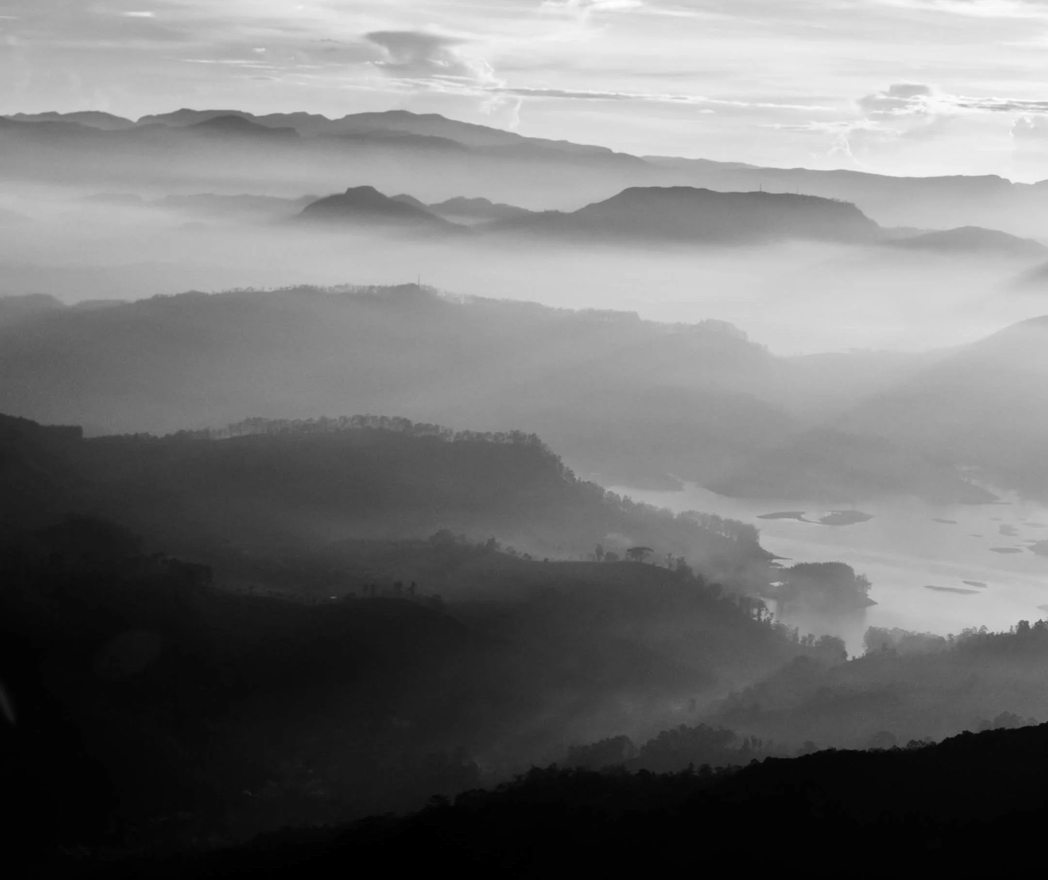 Black and white photograph of mountain layers with fog and water bodies in the valleys.