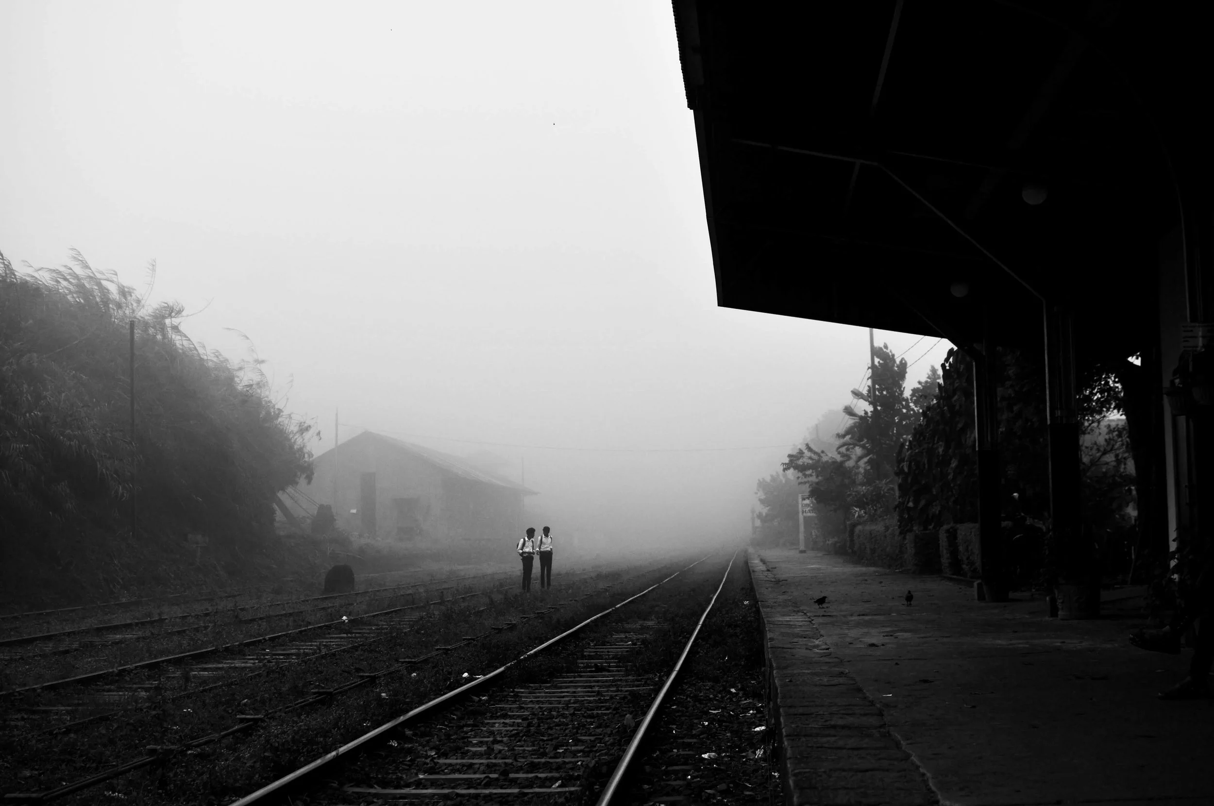 Two people with backpacks standing on a foggy train track at a small train station, with a building and trees in the background.