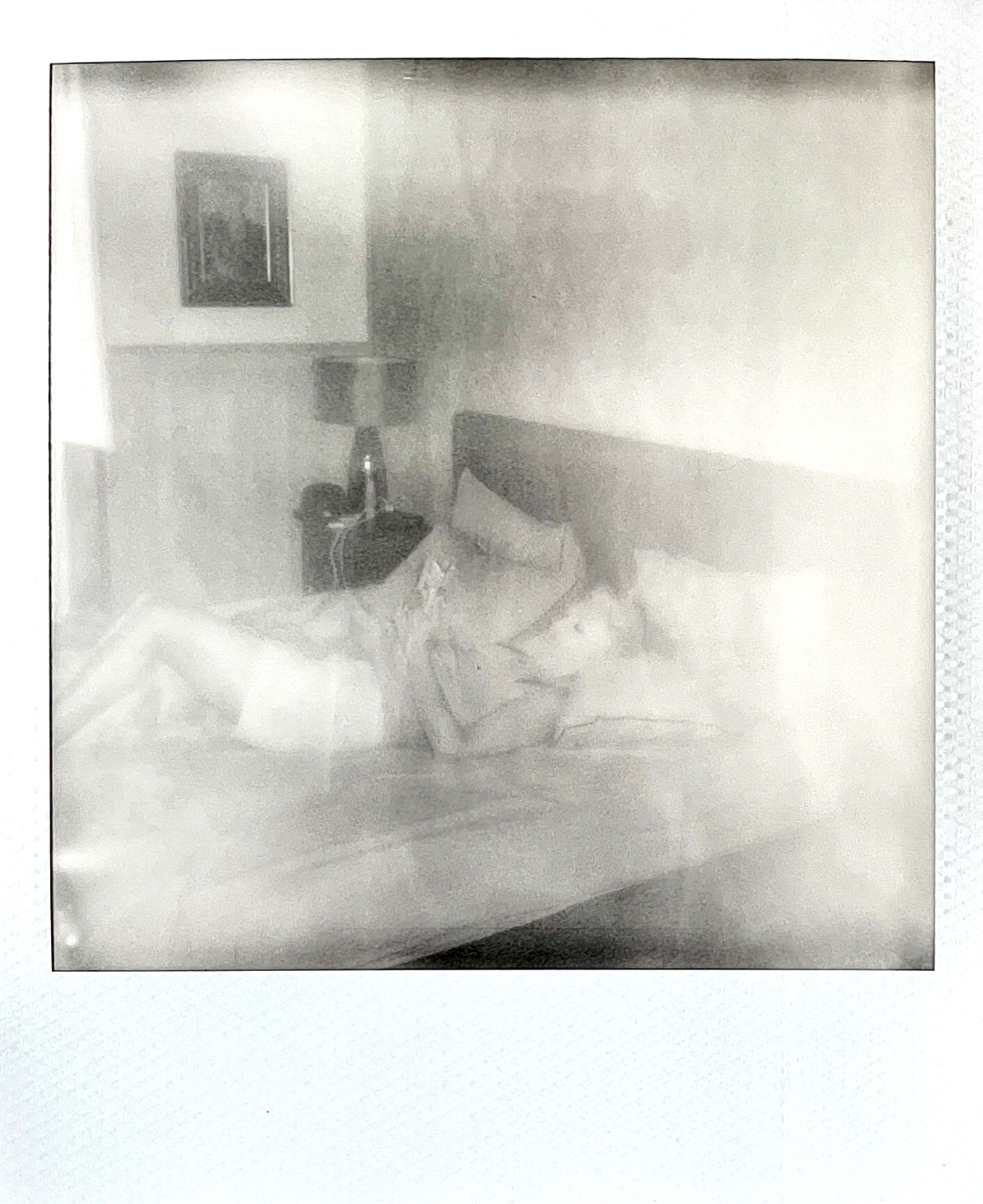 A person sleeping on a bed in a hotel room, with a nightstand, lamp, and framed picture on the wall.