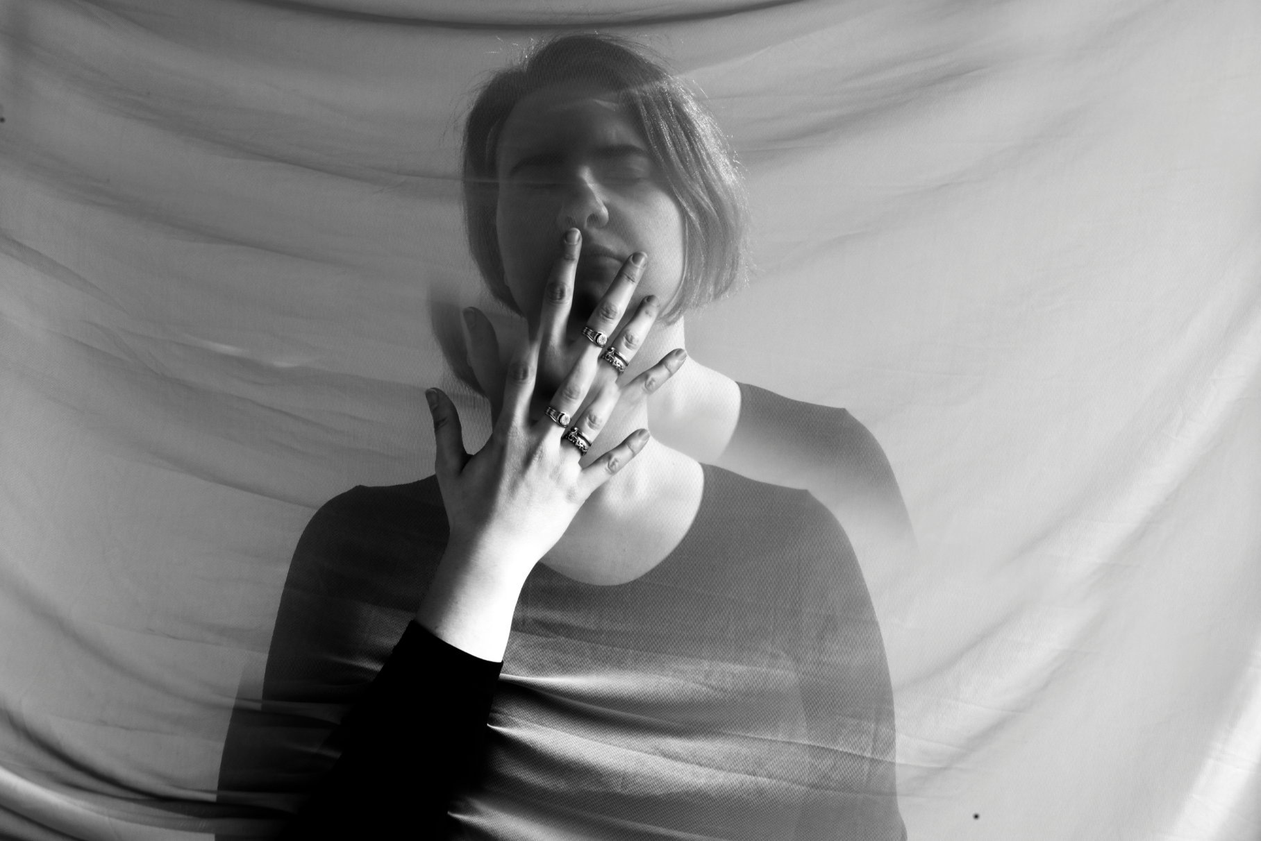 A young woman with short hair and wearing rings on her fingers touches her lips with her hand. She is seen through a semi-transparent fabric, with her face softly illuminated against a light background.
