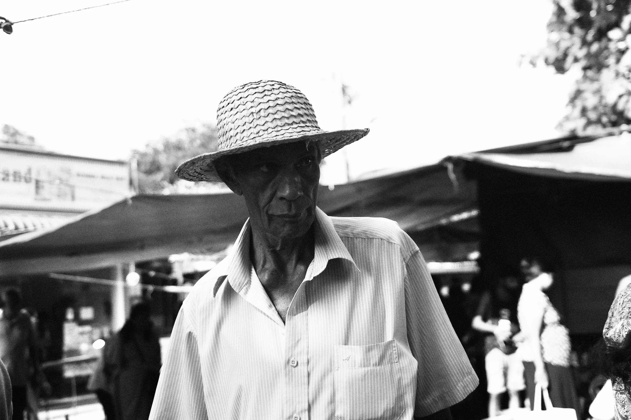A man wearing a straw hat and a striped short-sleeve shirt at an outdoor market, with blurred stalls and people in the background.