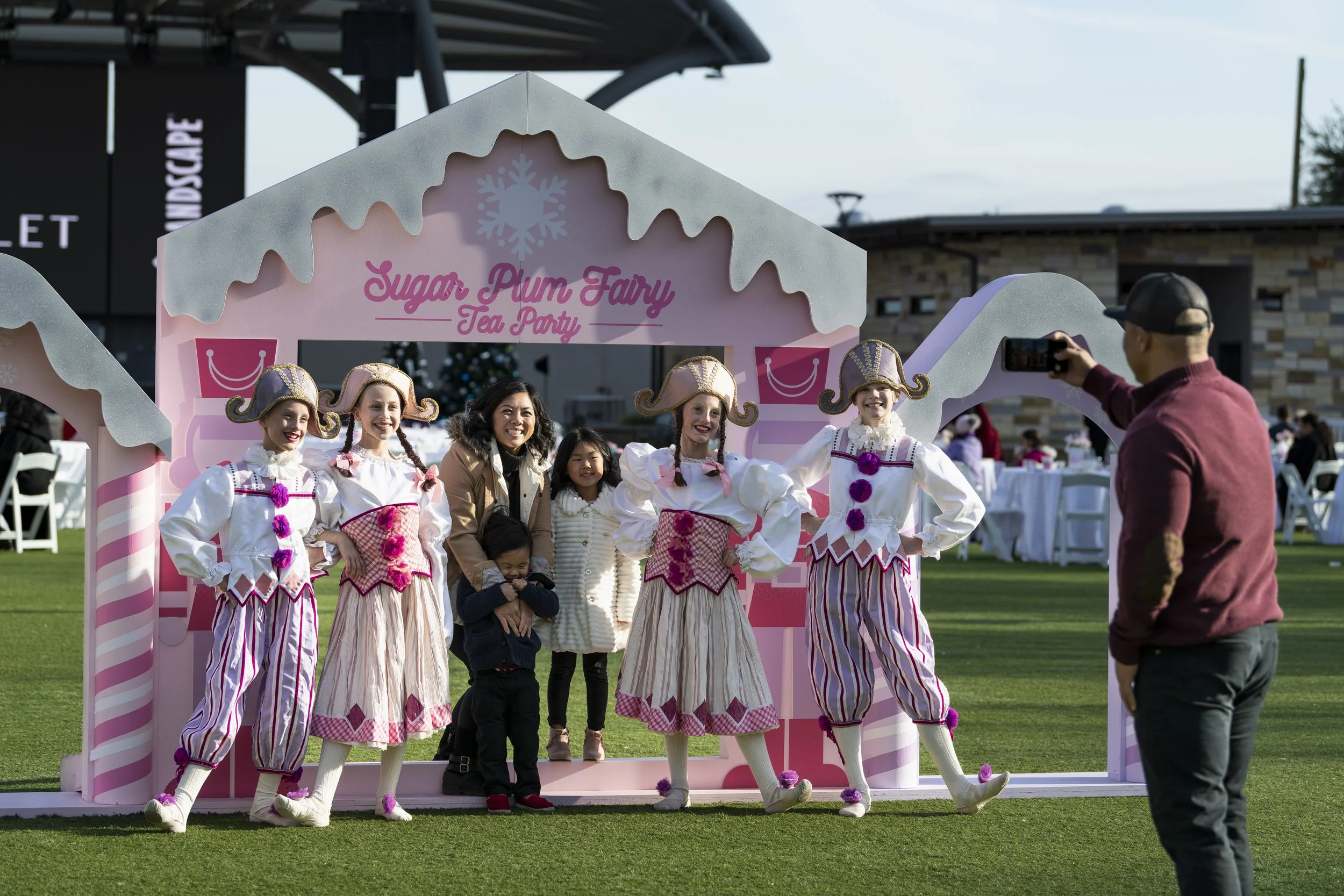 A group of children dressed as sugar plum fairies in pink and white costumes posing for a photo at a tea party event with an adult woman and two young children standing behind them, outside on a grassy area, with a man taking their photo.