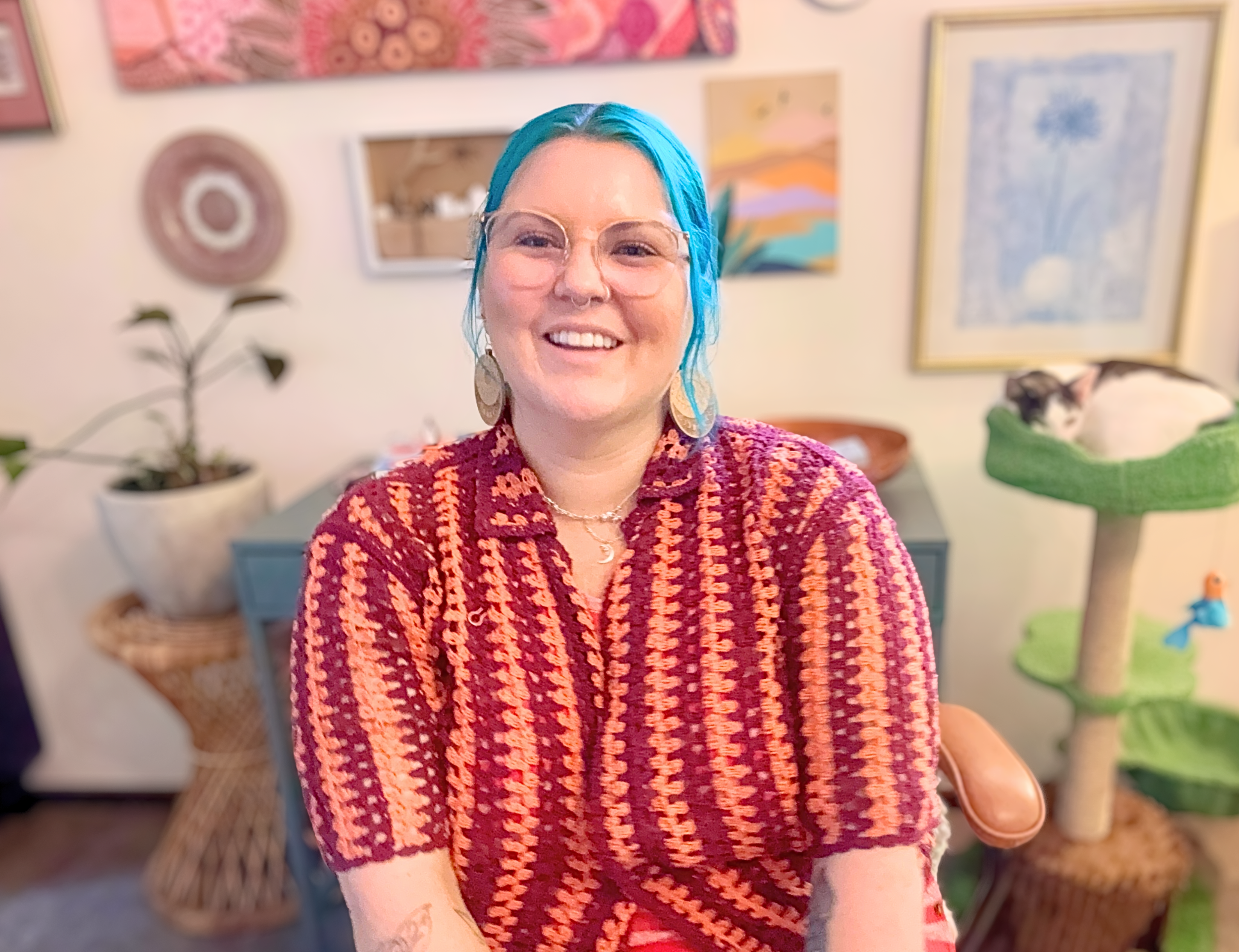 Person with blue hair, glasses, crochet top, smiling and in the background there is a cat, plant, pictures.