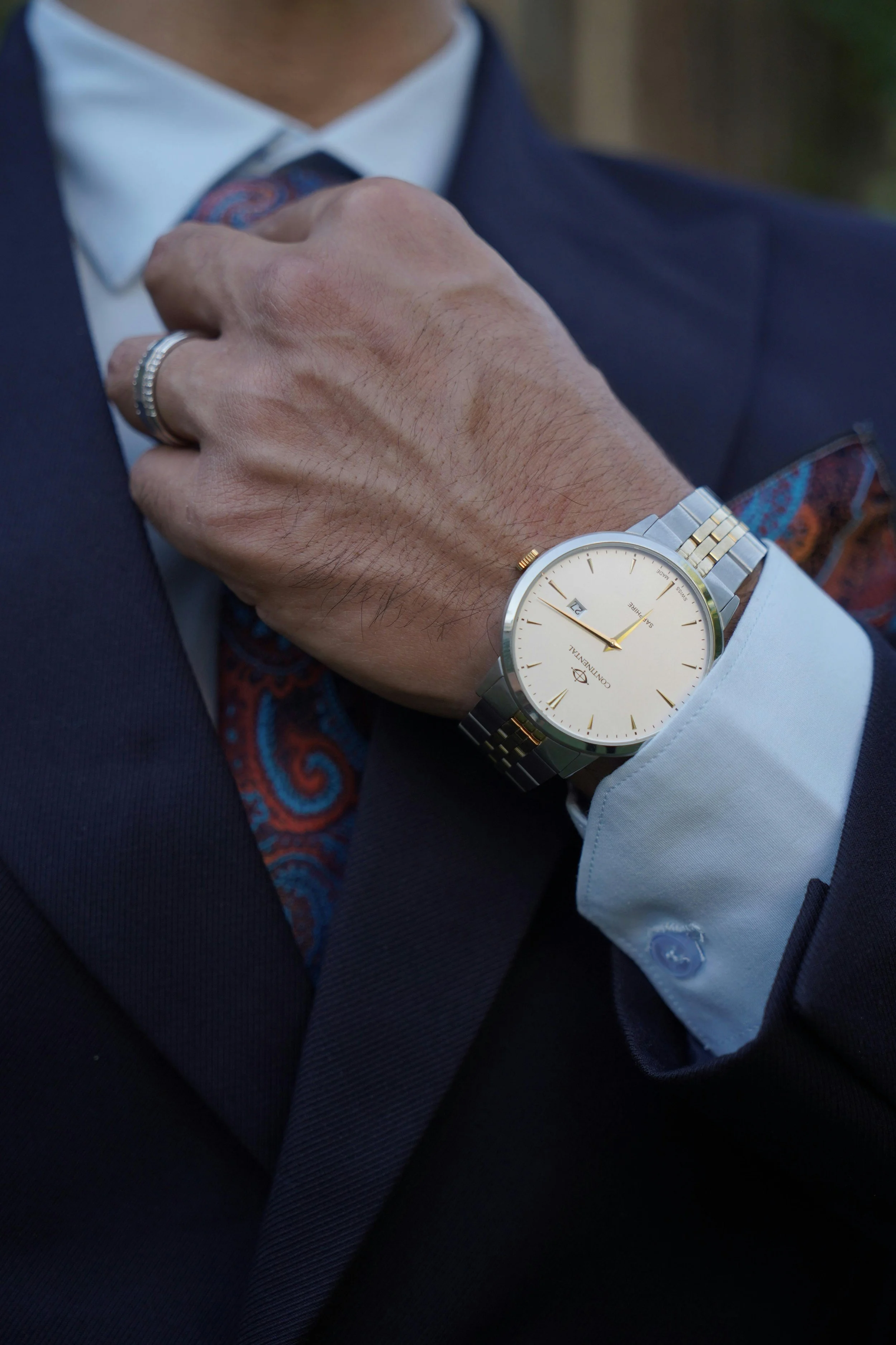 Close-up of a man's wrist wearing a silver and gold watch with a white face, dressed in a navy suit, white shirt, and patterned tie, adjusting his collar.