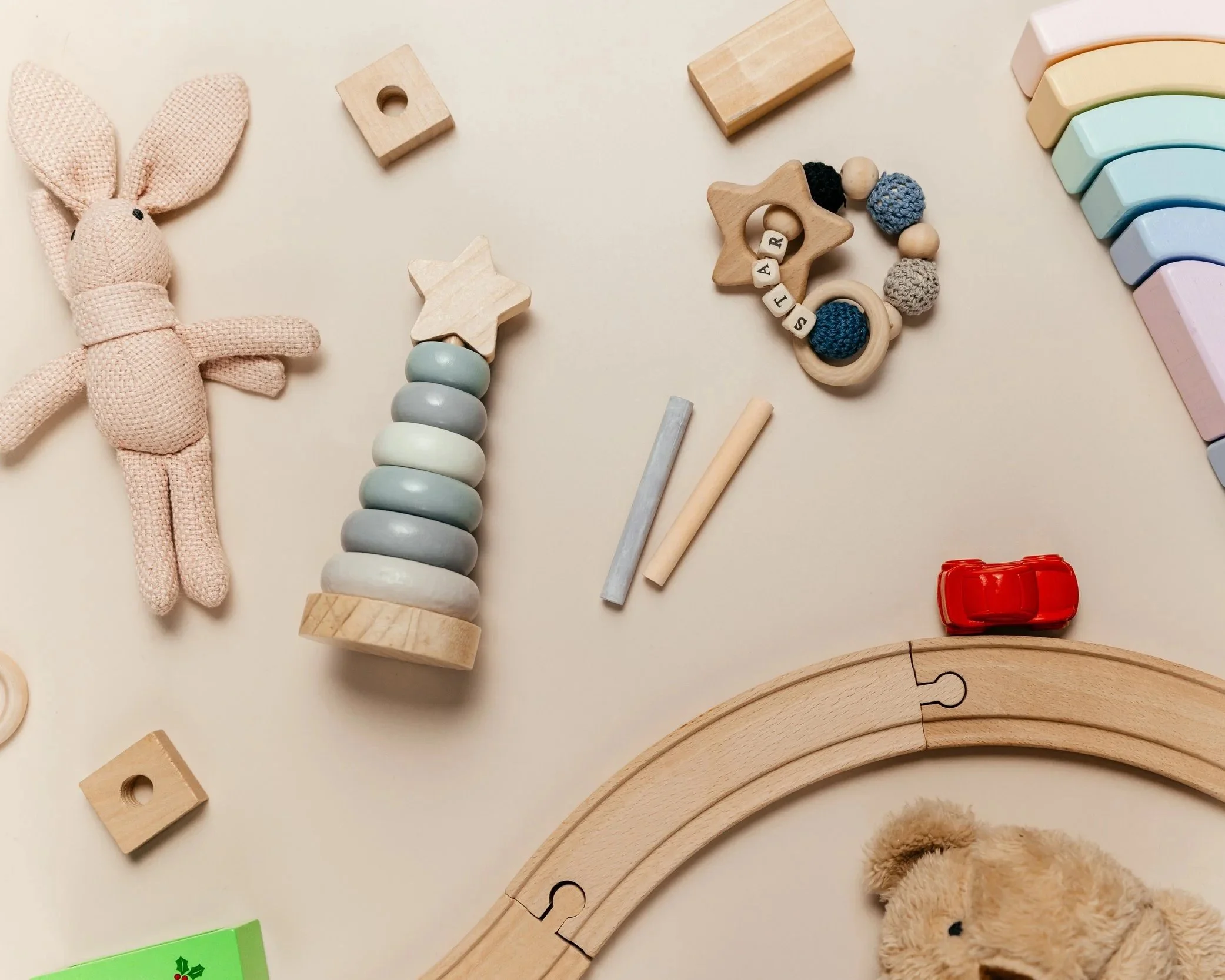 Children's toys on a beige surface, including a pink fabric bunny, a stack of pastel-colored wooden rings, a wooden star-shaped rattle with beads spelling "JAR," a red toy car, colorful semi-circular wooden blocks, and a curved wooden track with a teddy bear.