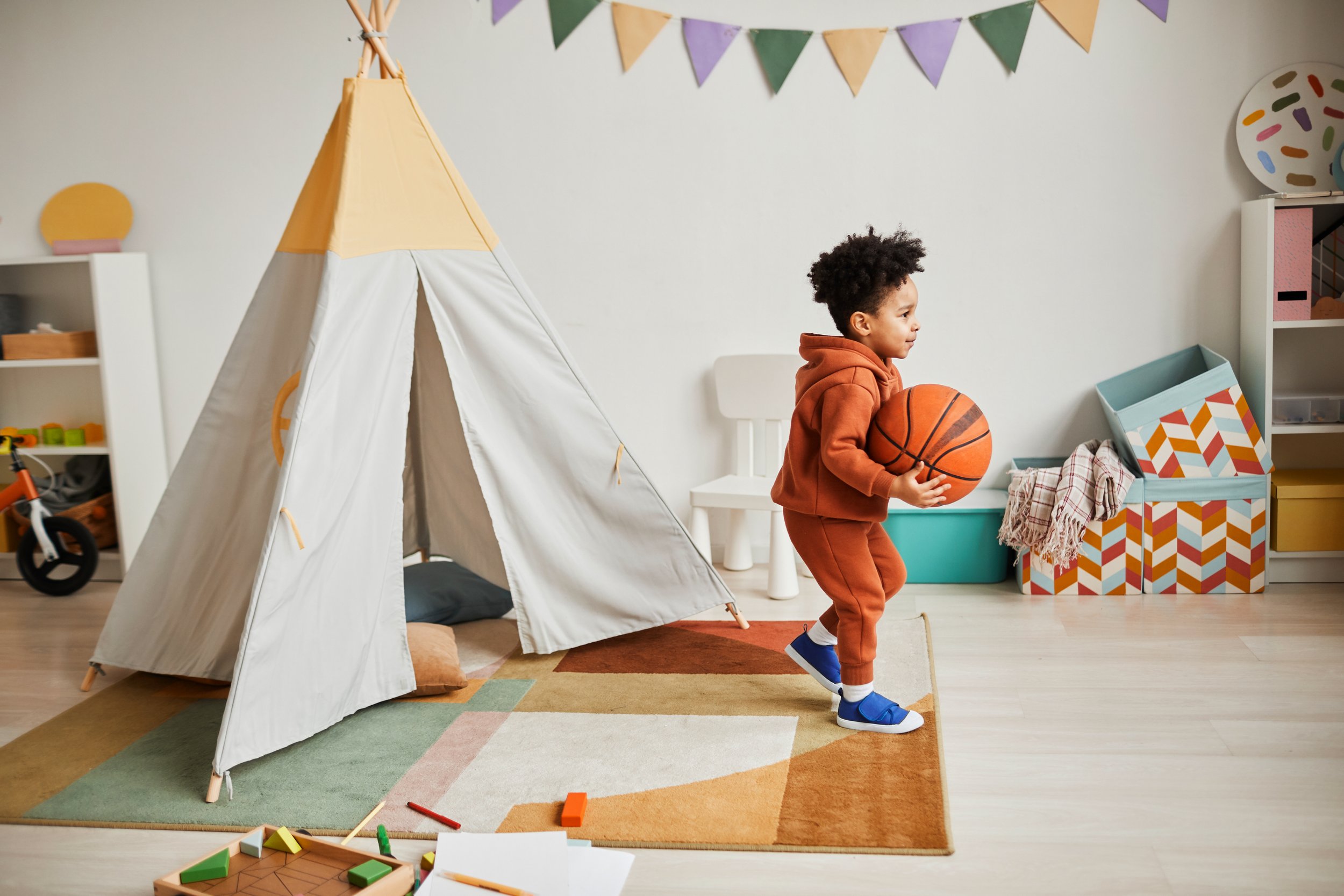 Young boy in orange hoodie and sweatpants holding a basketball in a playroom with a teepee tent, colorful bunting, and shelves with toys and storage boxes.