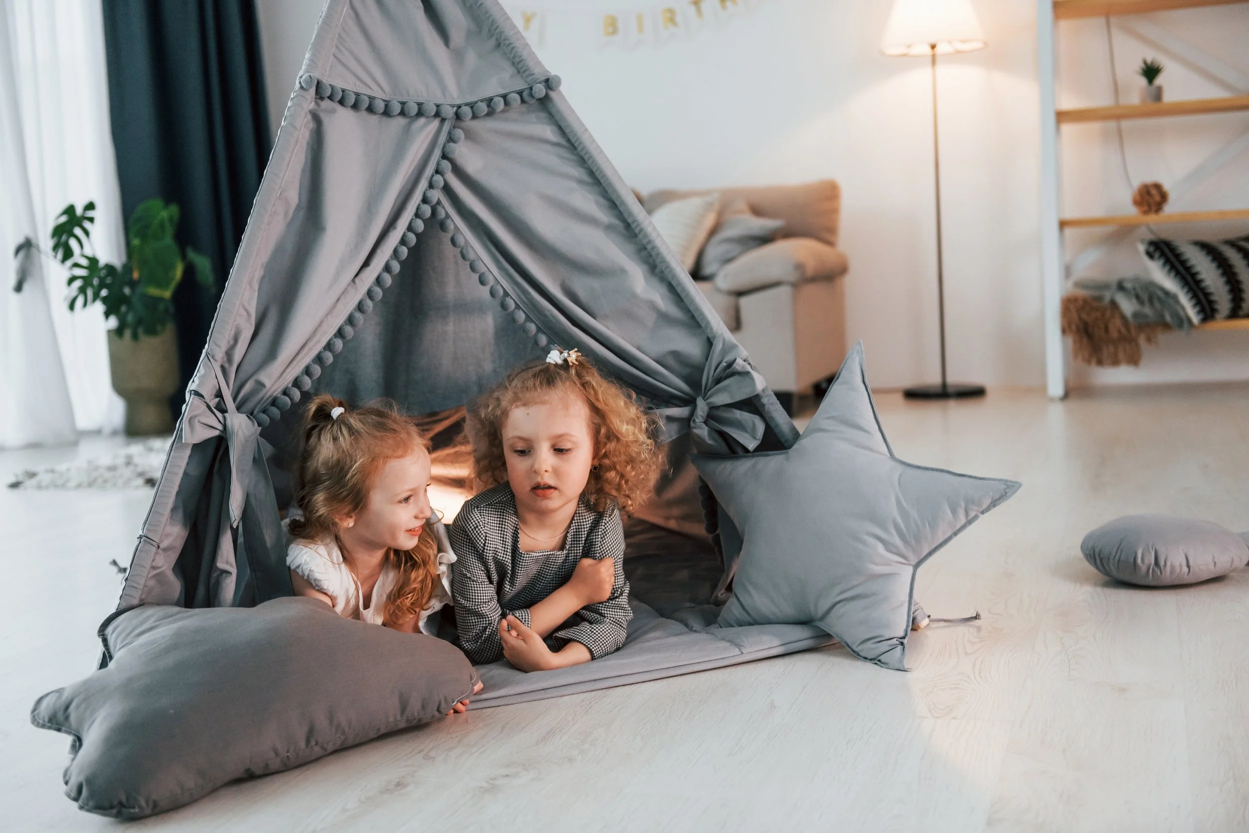Two young girls with light skin and curly hair are inside a cozy gray fabric play tent shaped like a house, on a light-colored floor in a living room. The tent has star-shaped and round pillows nearby, with a sofa, bookshelf, and standing lamp in the background.